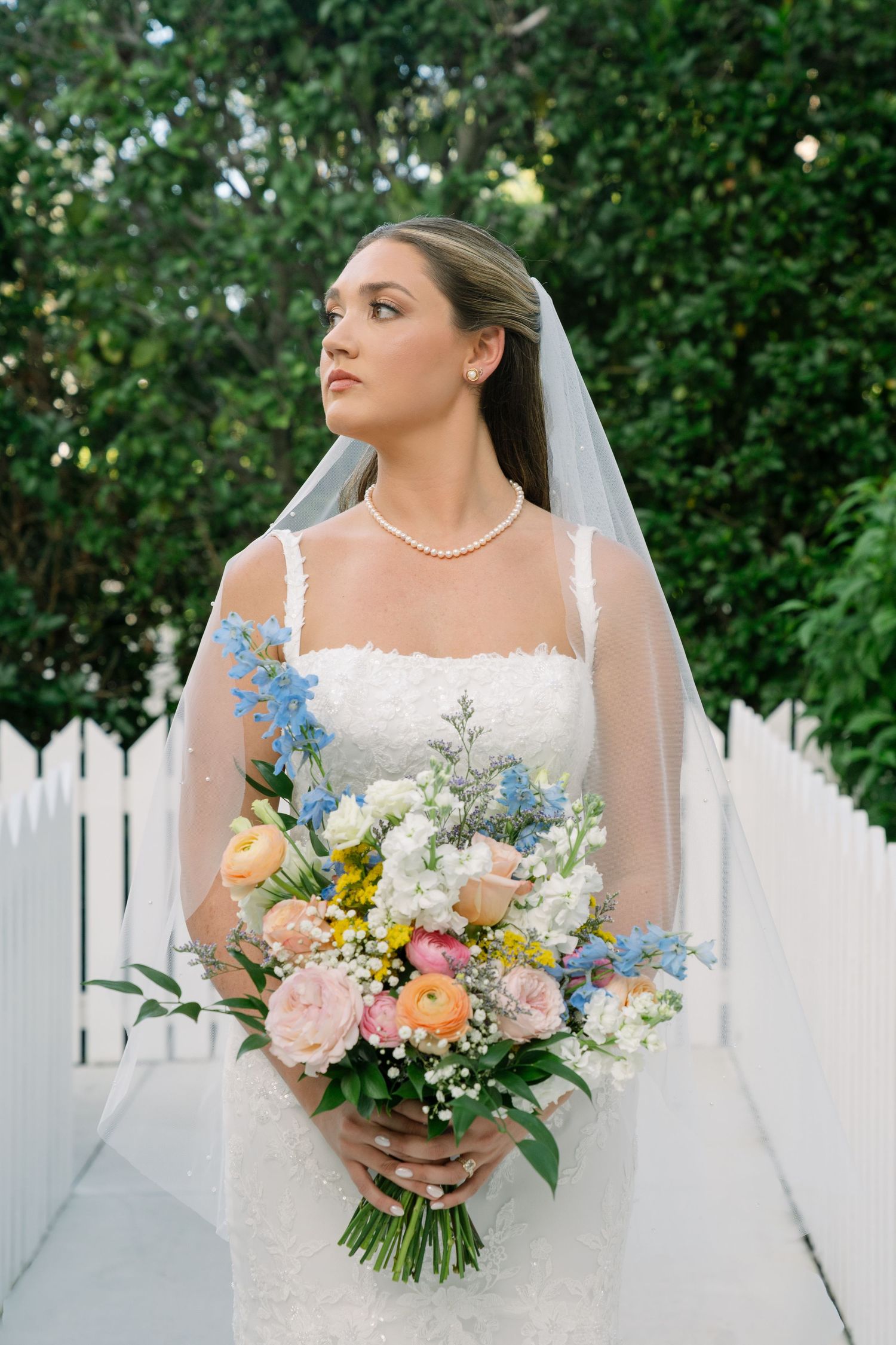 Beautiful bridal portrait with a lush bouquet of peach, blue and white flowers against a white picket fence backdrop.