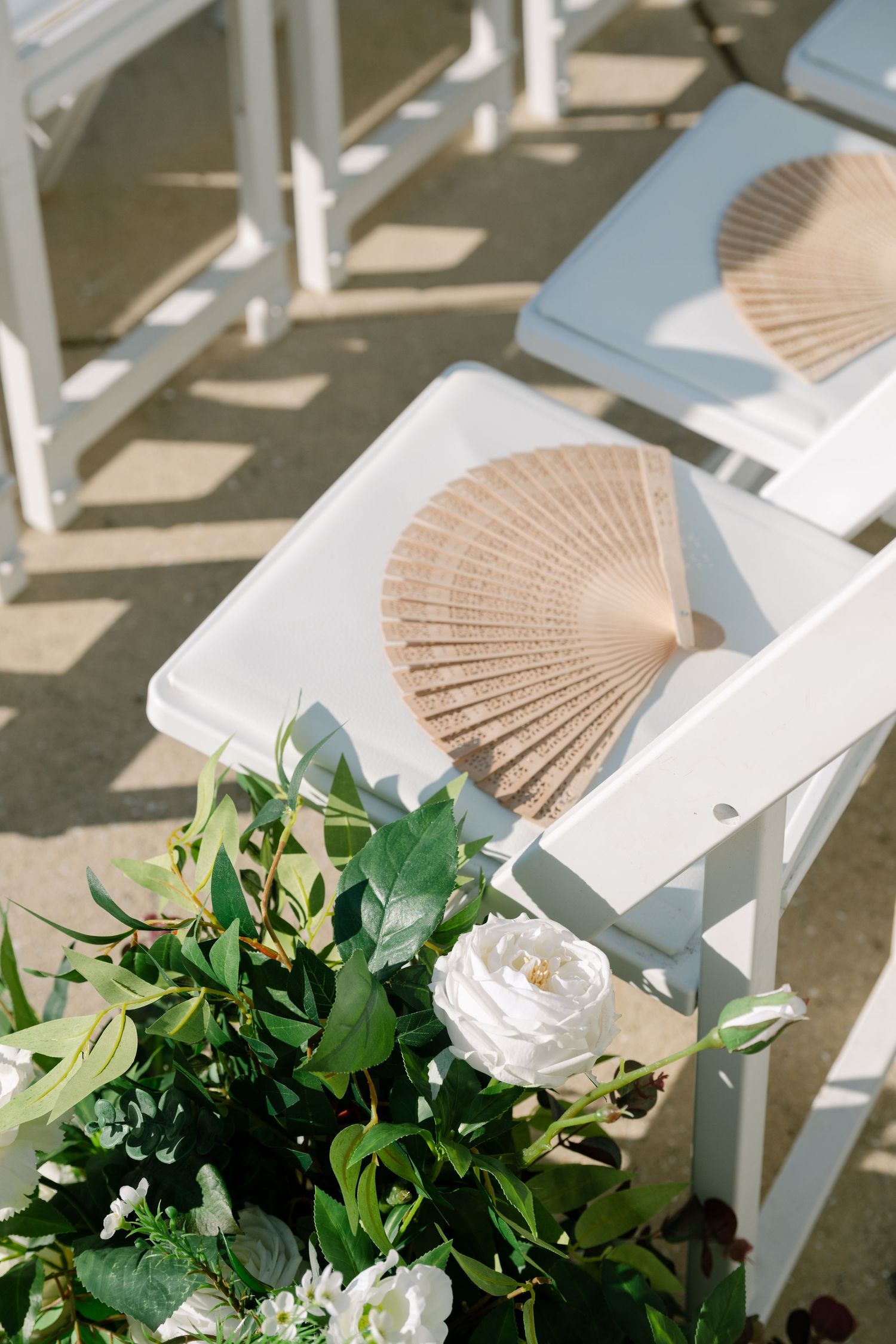 White wooden ceremony chairs decorated with floral arrangements and tan paper fans.