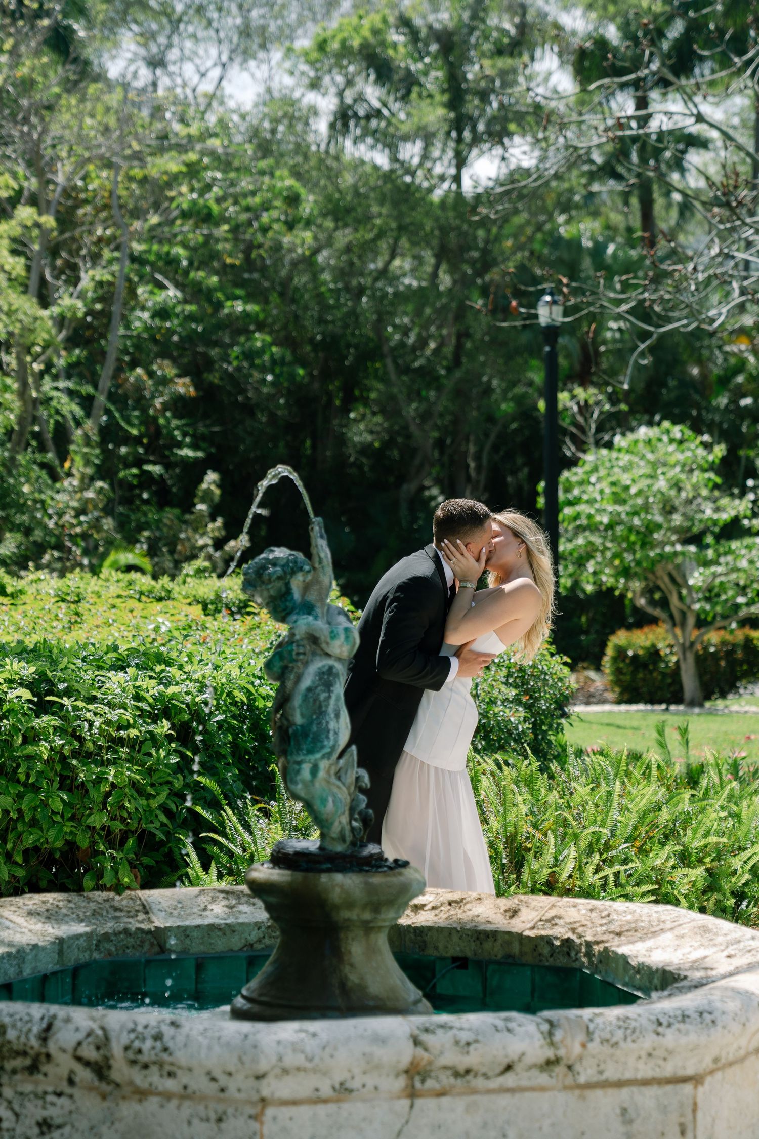 Bride and groom sharing a romantic kiss beside a cherub fountain in a lush green garden setting.