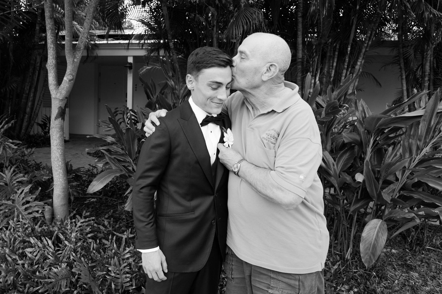 Older man kisses groom on cheek in black and white wedding photo outdoors.
