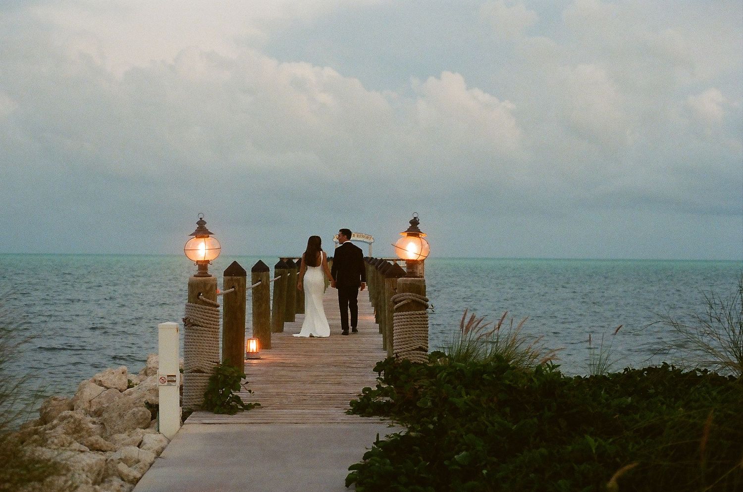Couple in wedding attire standing on a wooden dock overlooking the ocean at dusk with glowing lanterns.