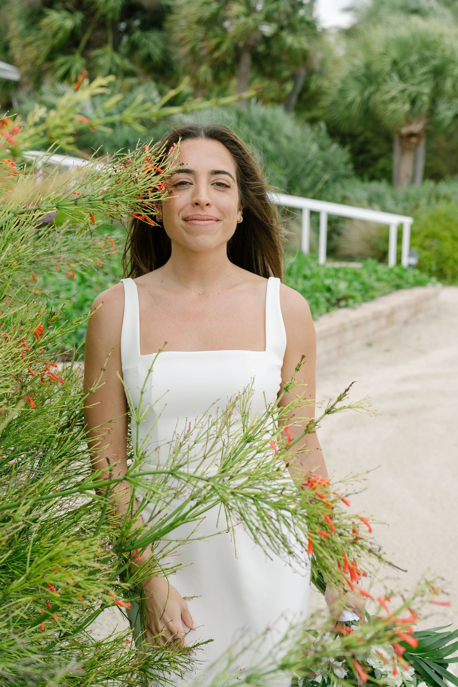 Bride in white sleeveless dress standing among vibrant green plants with red flowers.