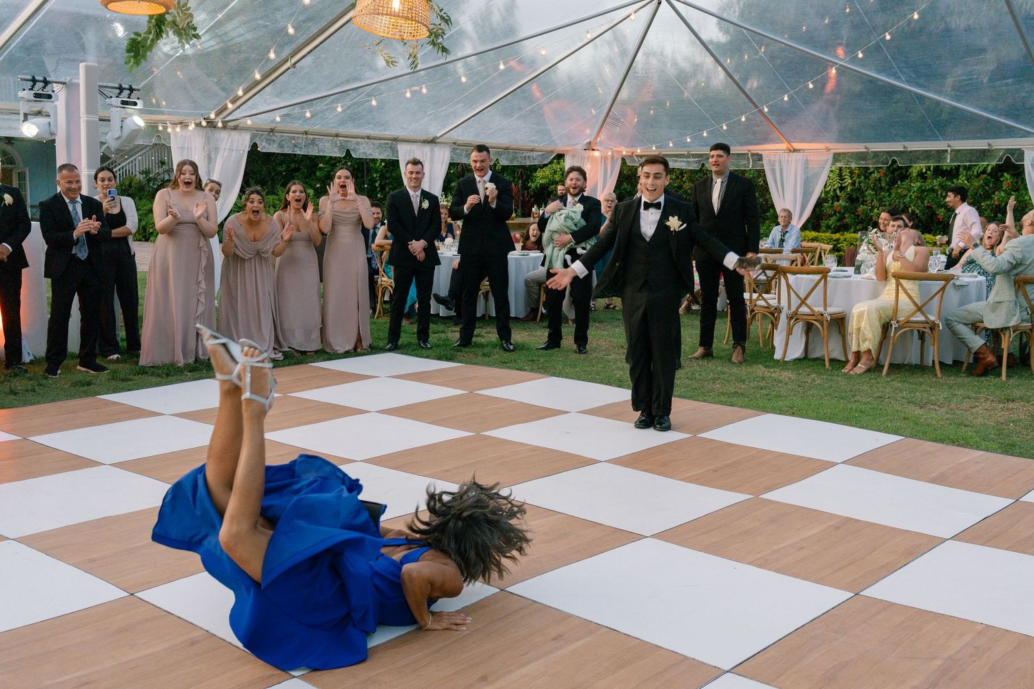 Energetic wedding performer in a blue dress does a floor move on a checkered dance floor at a reception.