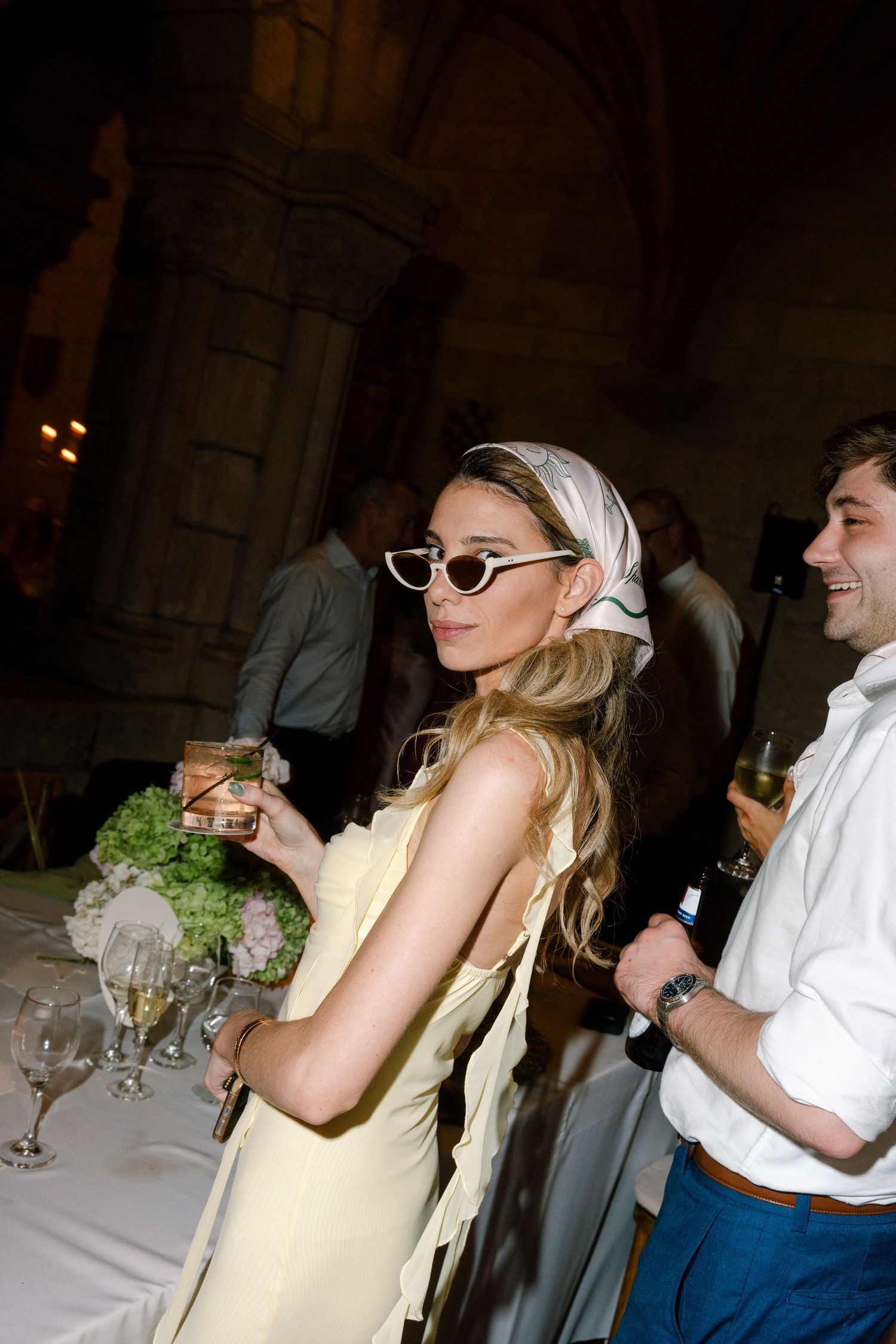 Stylish wedding guest in yellow dress, cat-eye sunglasses and silk headscarf holding drinks at evening reception.