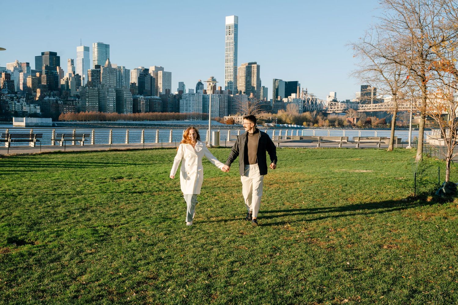 Two people walk their dogs in a grassy park with Manhattan skyline views on a sunny autumn afternoon.