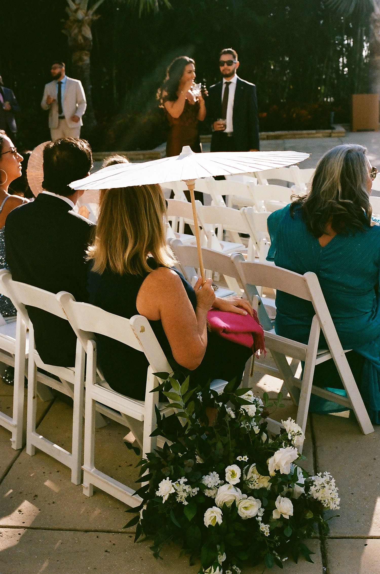White folding chairs with floral arrangements line an outdoor wedding ceremony venue.
