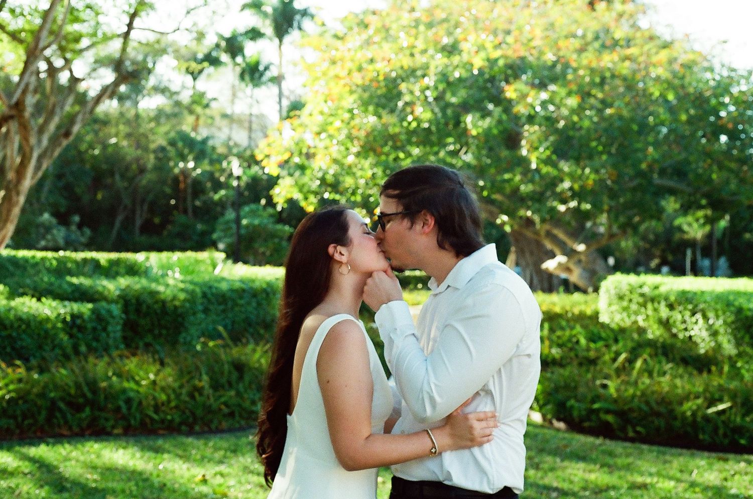 A romantic couple shares a tender kiss in a lush tropical garden with flowering yellow trees in the background.