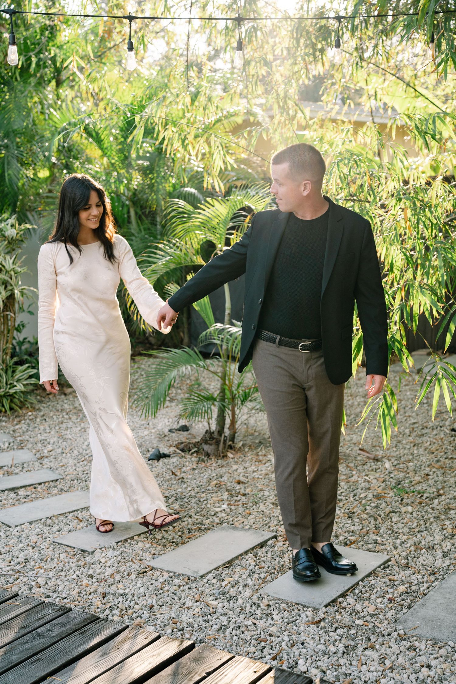 A couple stands together on a gravel path surrounded by lush bamboo plants in an outdoor garden setting.
