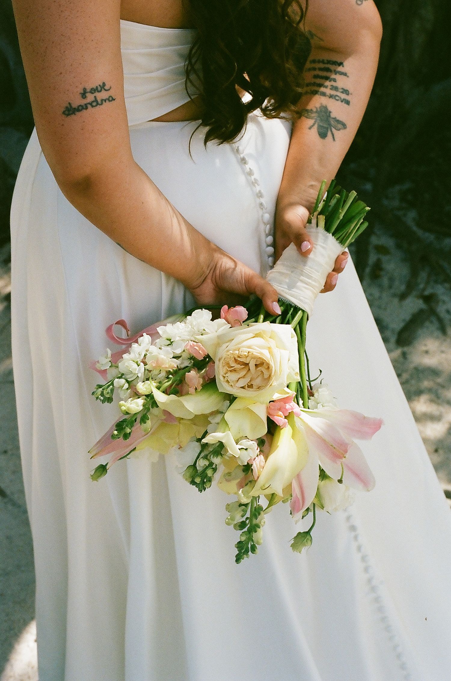 A bride holding a bouquet of flowers with a floral tattoo visible on her arm.
