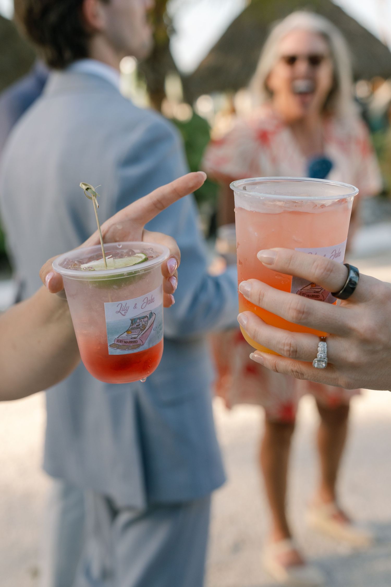 The image shows two people holding colorful cocktail drinks at an outdoor event.