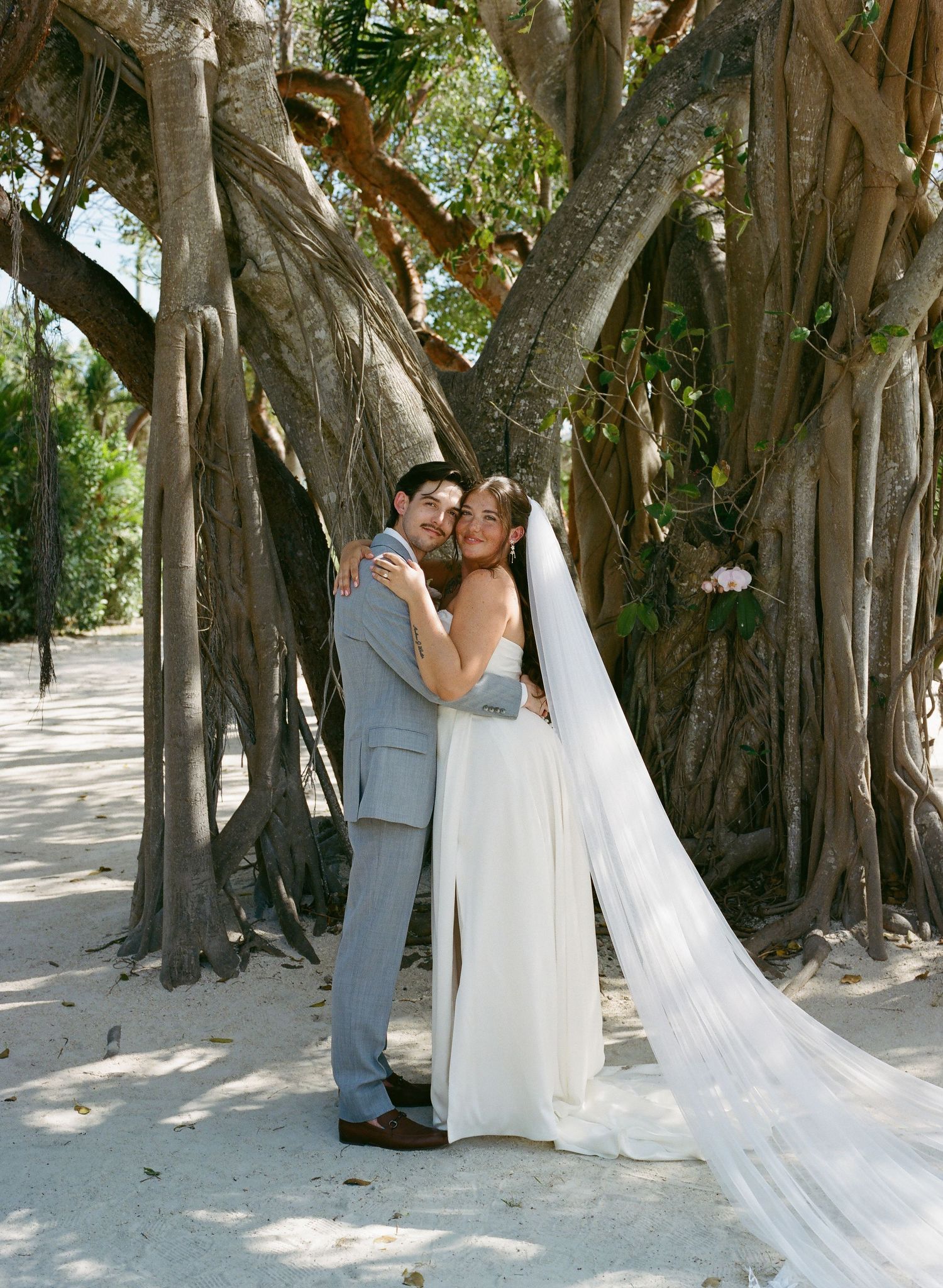 Bride in white gown with veil and groom in gray suit embrace under tropical banyan tree.