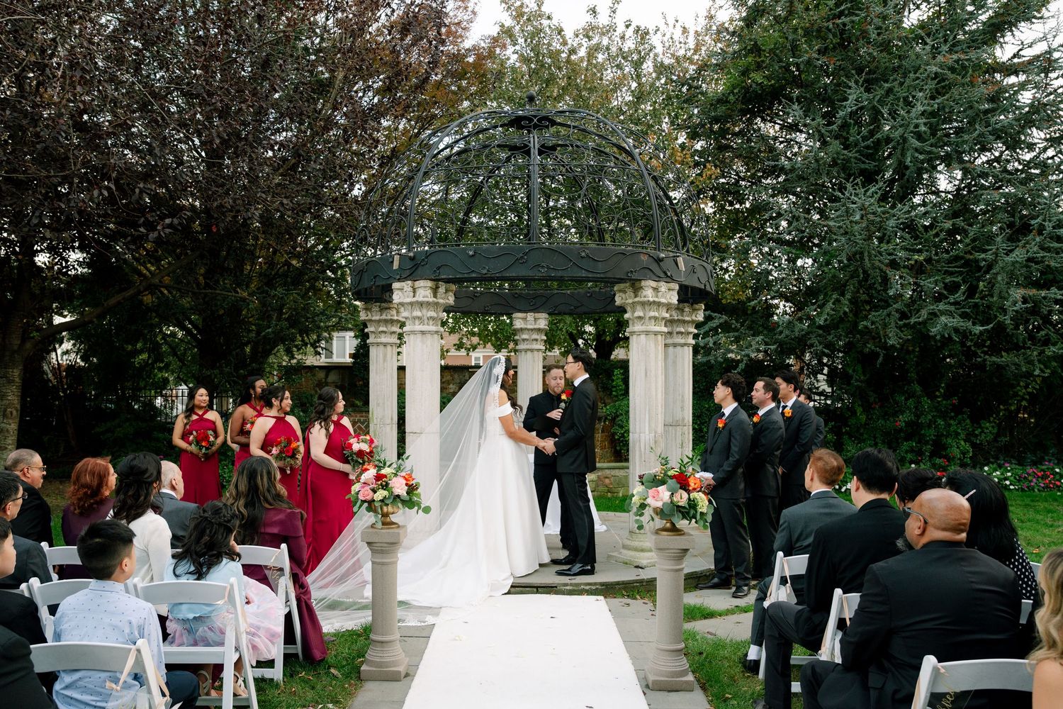 Outdoor wedding ceremony takes place under an elegant stone gazebo with red-clad bridesmaids and seated guests.