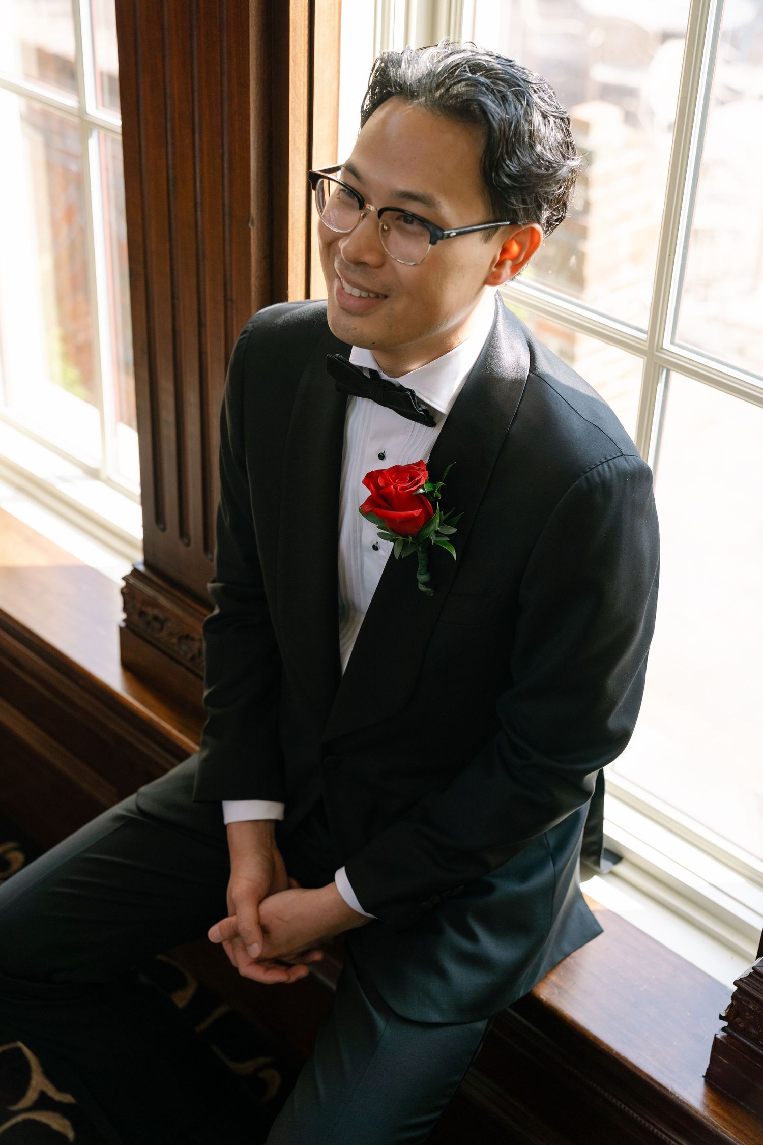 A groom in a black tuxedo sits by a window with natural light streaming in.