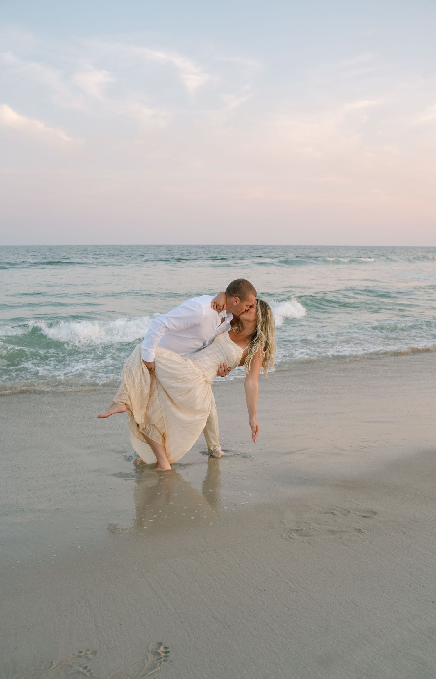Couple in white outfits playfully writing in the sand together at sunset on a peaceful beach.