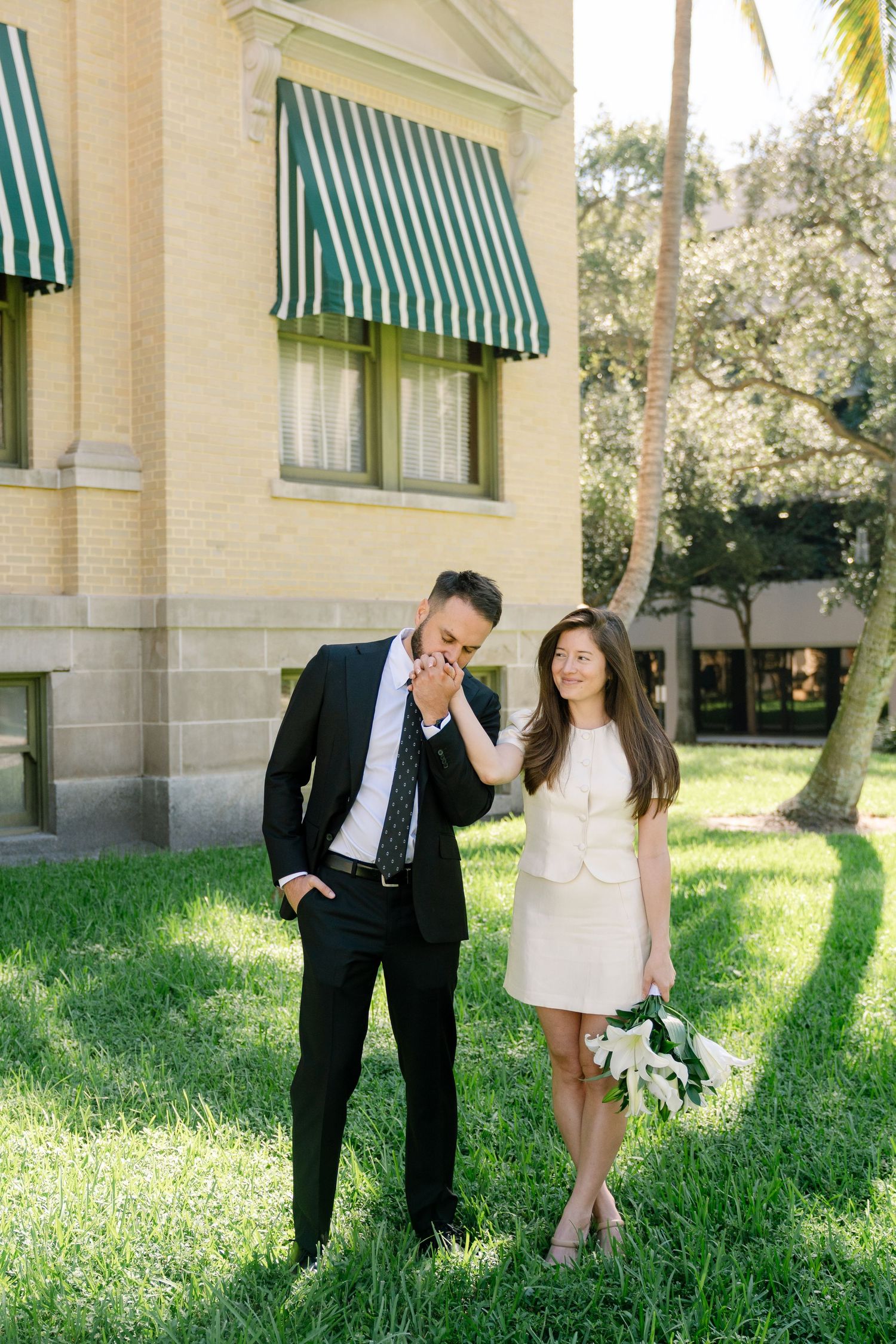 A couple poses playfully on a lawn in front of a building with green striped awnings.