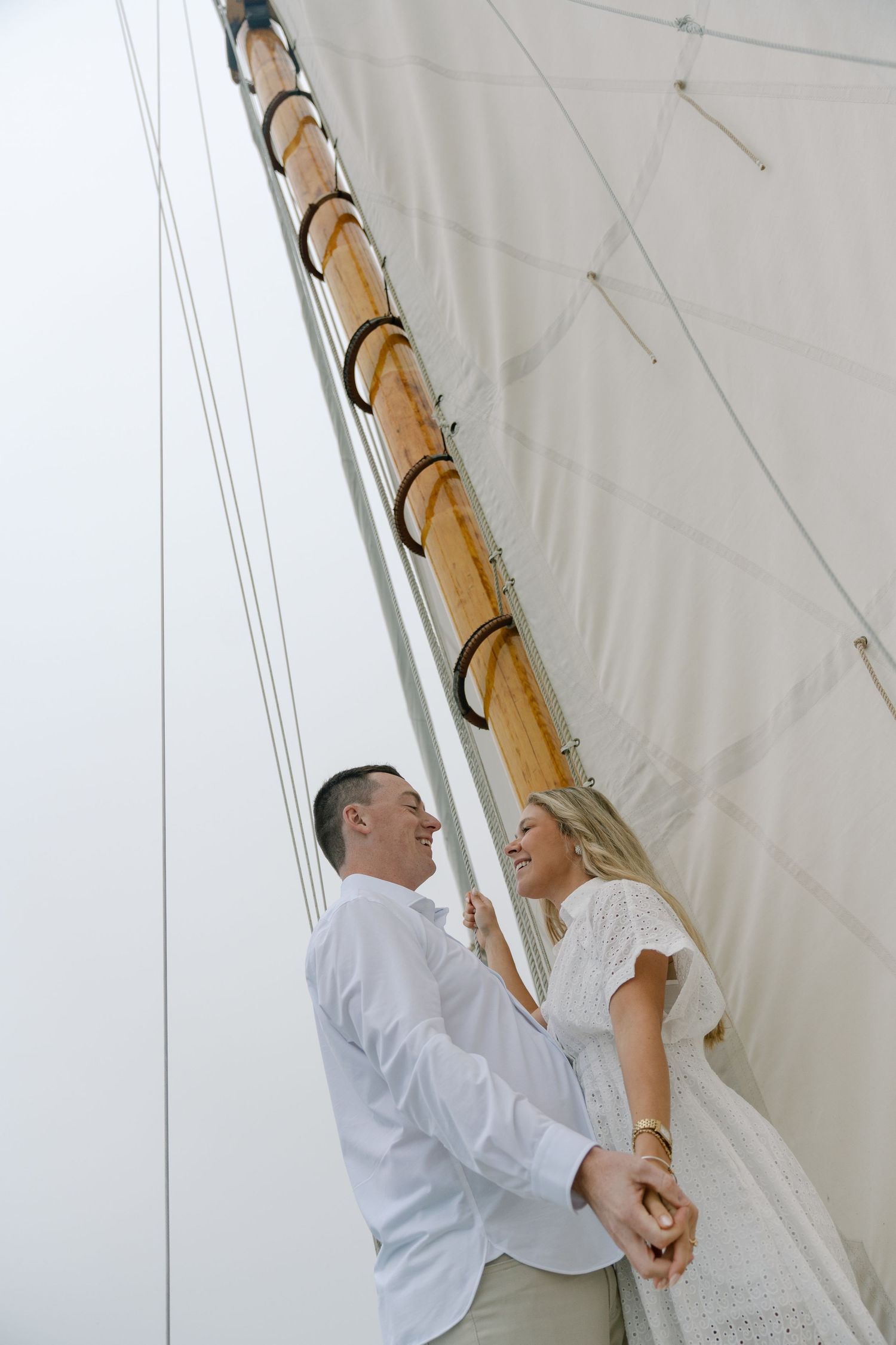 Couple in white outfits holding hands and smiling at each other beside a wooden mast and sail on a sailboat.