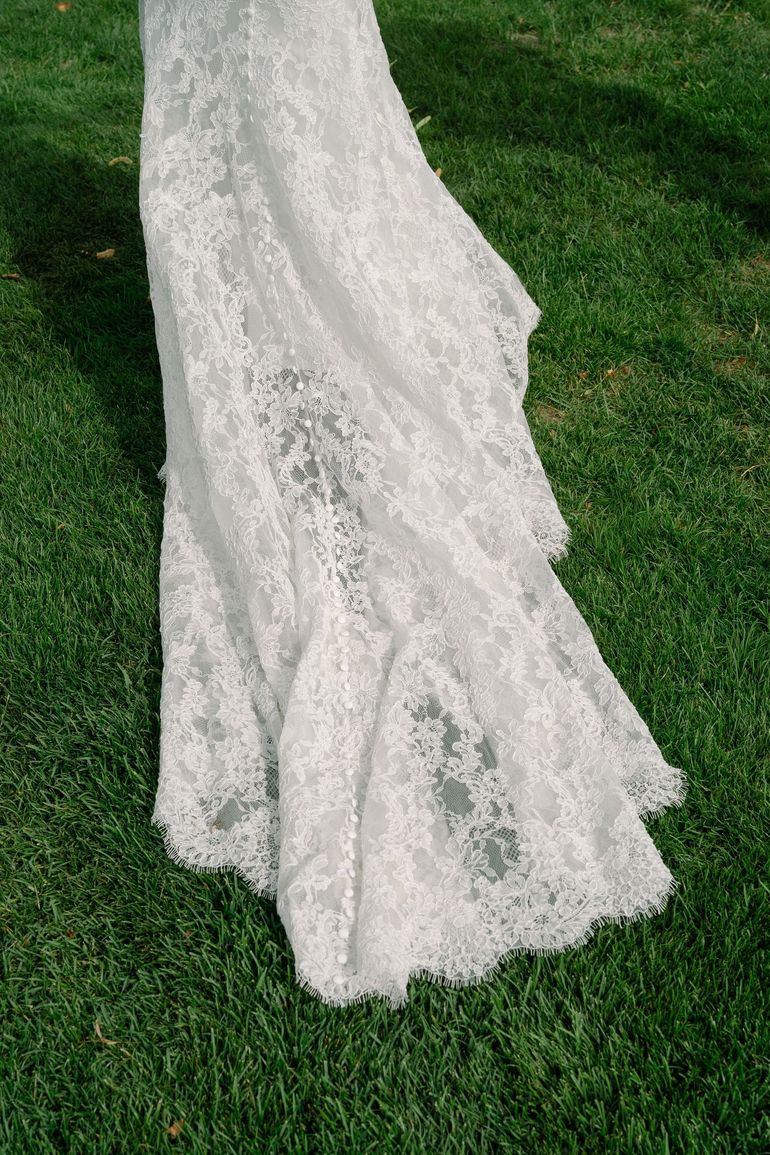 Close-up of a white lace wedding dress train spread across green grass outdoors.