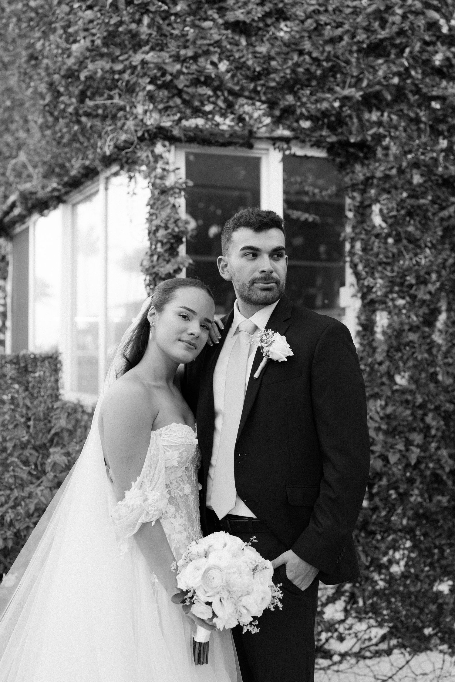 Black and white portrait of bride and groom posing before ivy-covered wall, bride holding white bouquet.