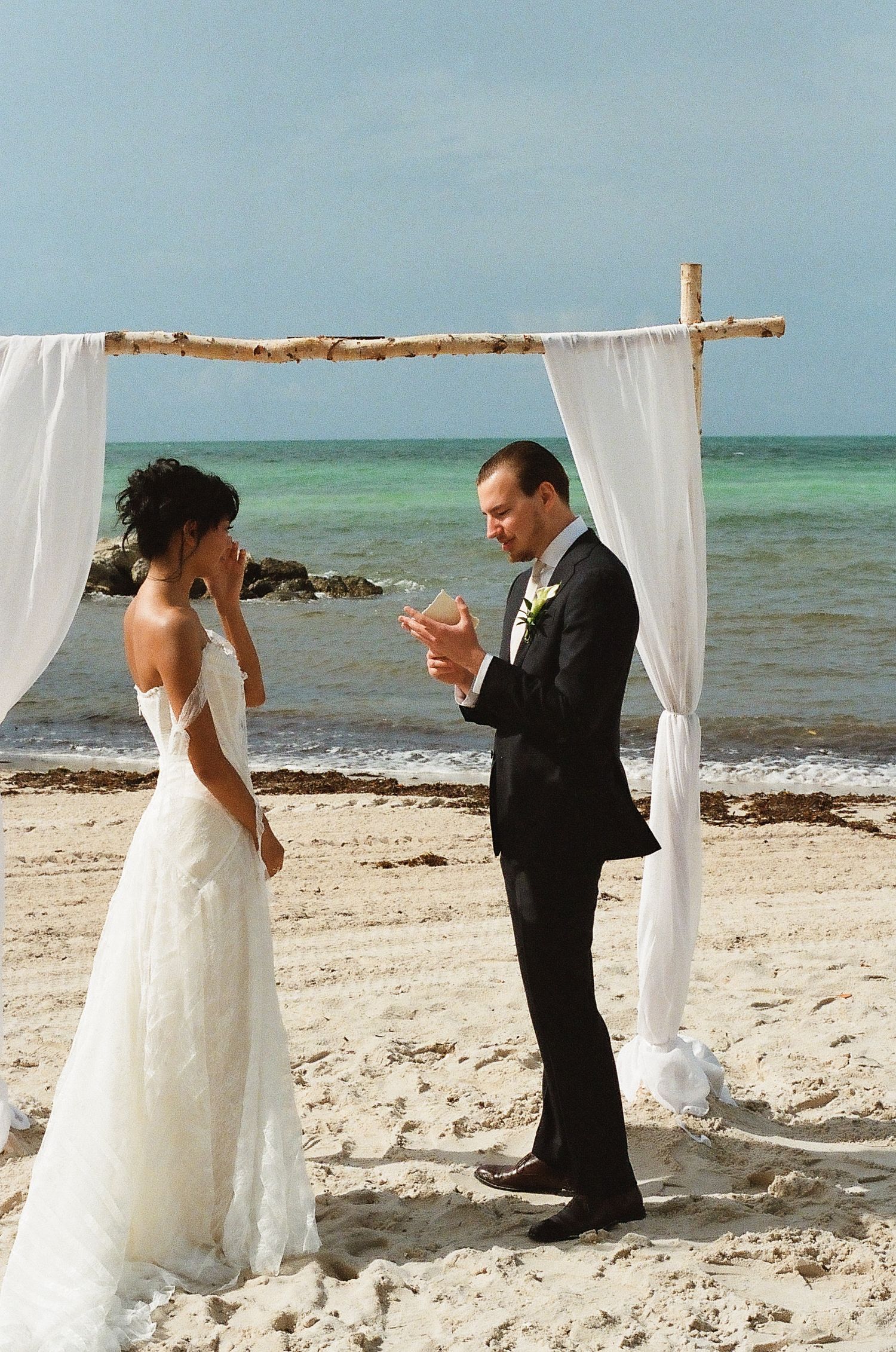 Couple exchanging vows at a romantic beach wedding ceremony under a white draped arch with turquoise ocean backdrop.