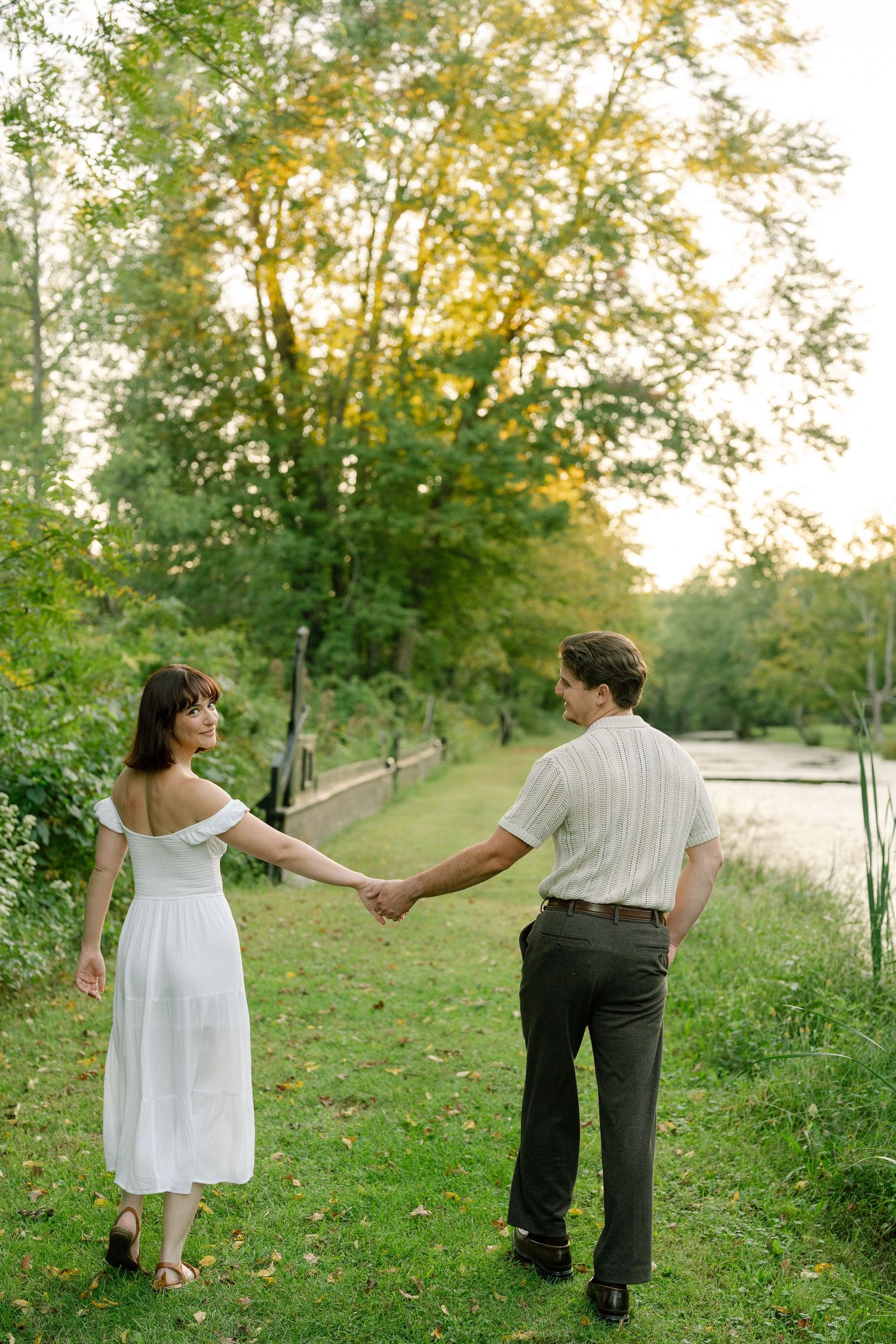 A couple holds hands while walking along a lush green riverbank at sunset.