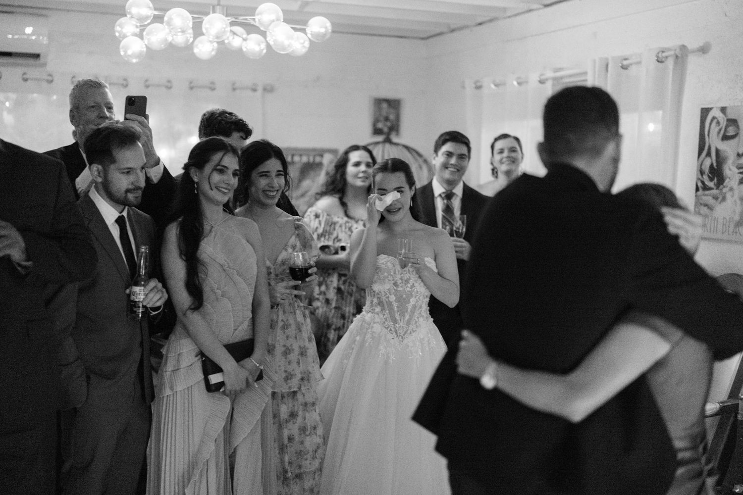 Black and white wedding photo of bride and groom sharing first dance while emotional guests watch and celebrate.