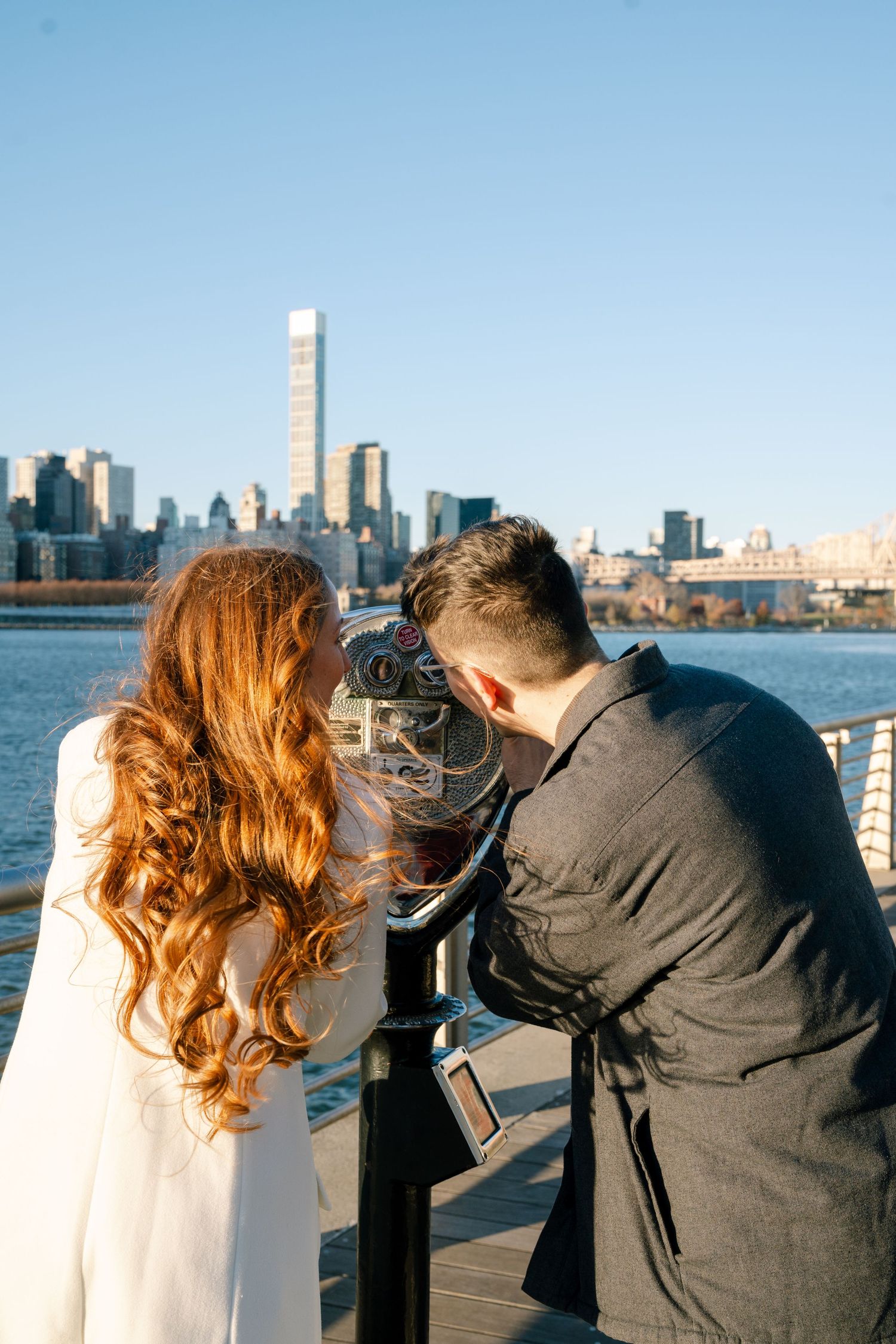 A couple looks through viewfinders at the New York City skyline across the water on a sunny day.