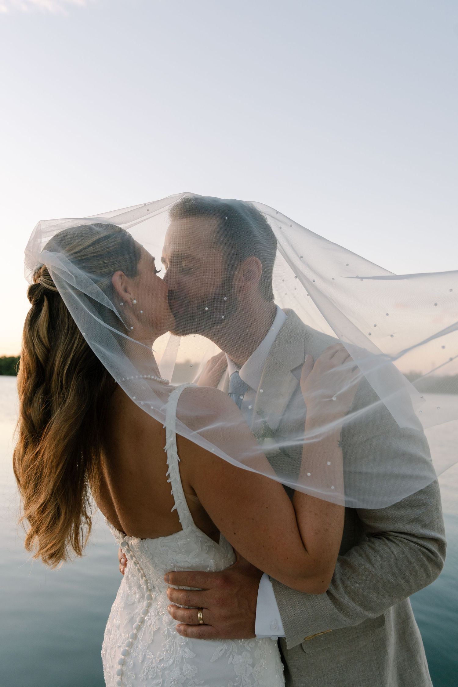 Romantic beach wedding photoshoot with couple kissing under flowing white veil at sunset.