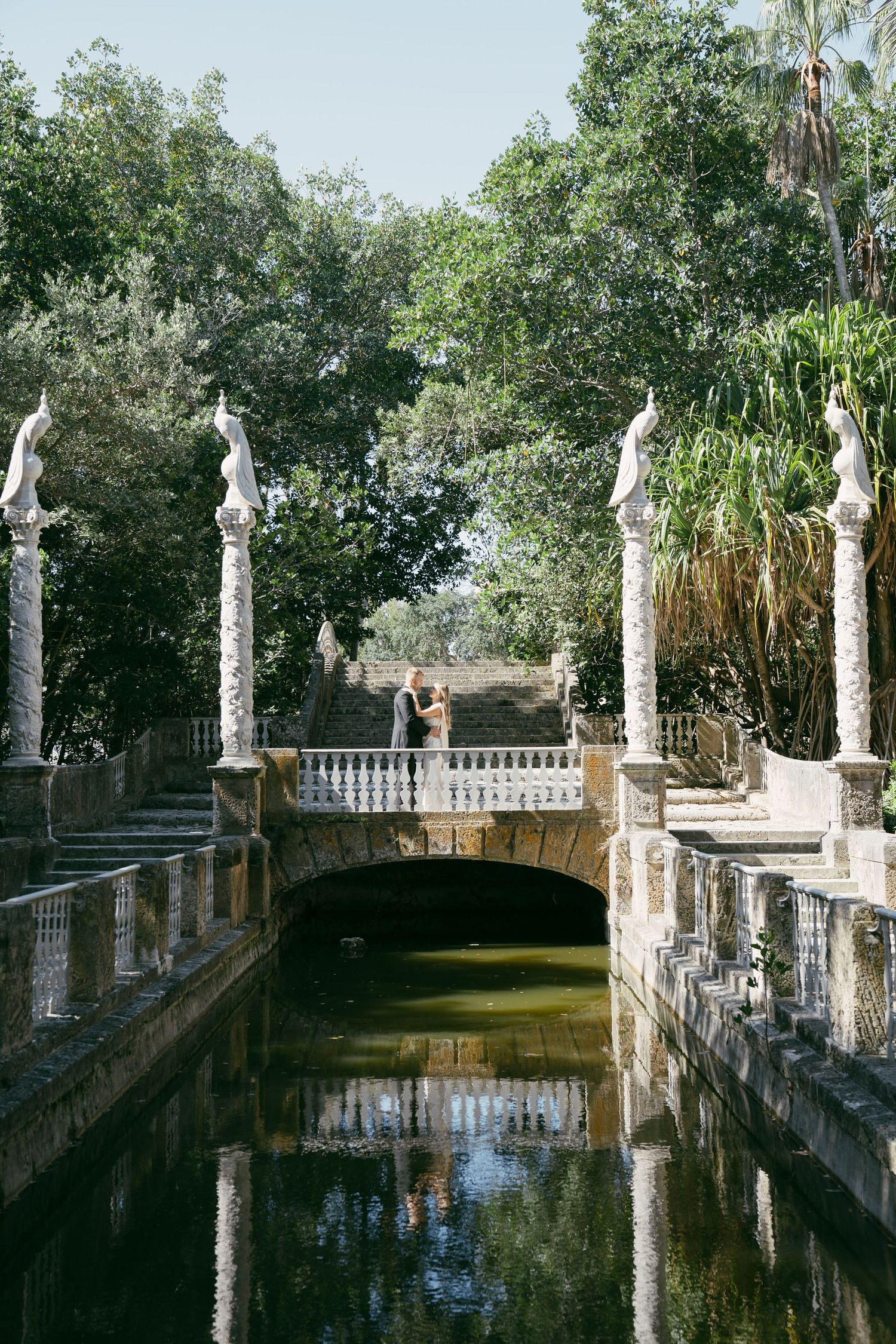 Ornate stone bridge with classical statues and balustrades reflects in a tranquil pool at Vizcaya Gardens in Miami.