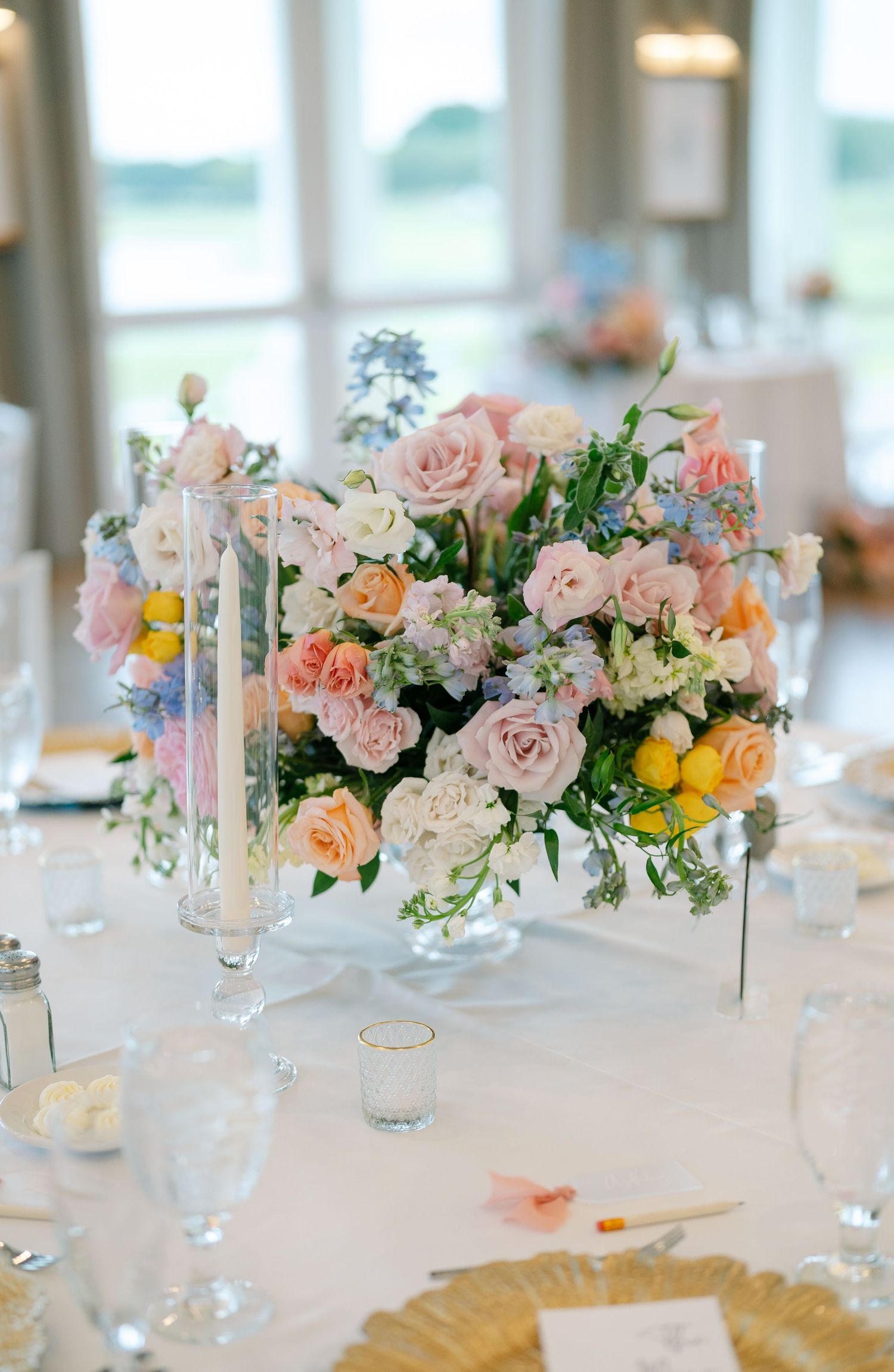 Elegant wedding centerpiece with soft pink roses, blue delphiniums, and yellow blooms arranged in white vase on formal table setting.
