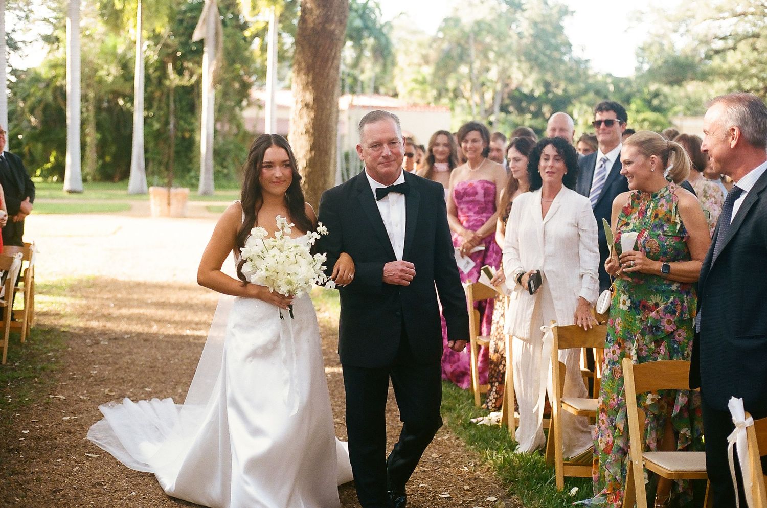 Bride in white gown walks down outdoor aisle with father in tuxedo, surrounded by wedding guests.