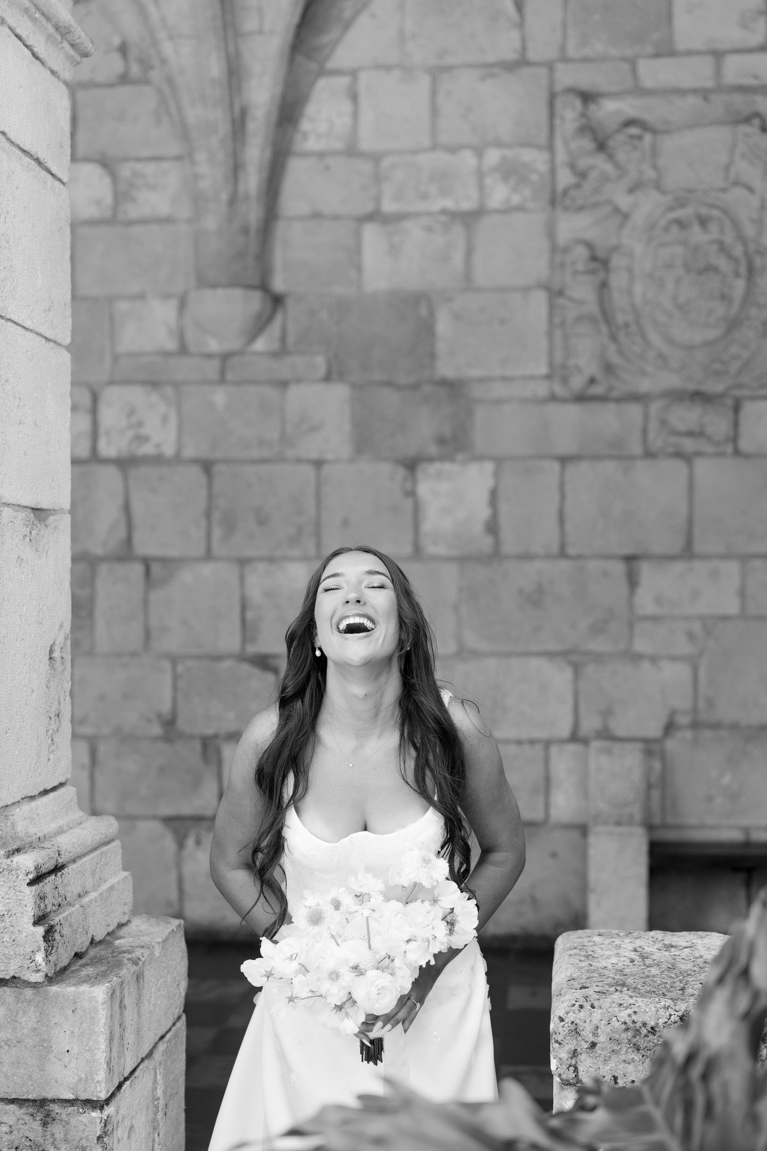 Black and white photo of a joyful bride in a wedding gown laughing while holding a floral bouquet outdoors.