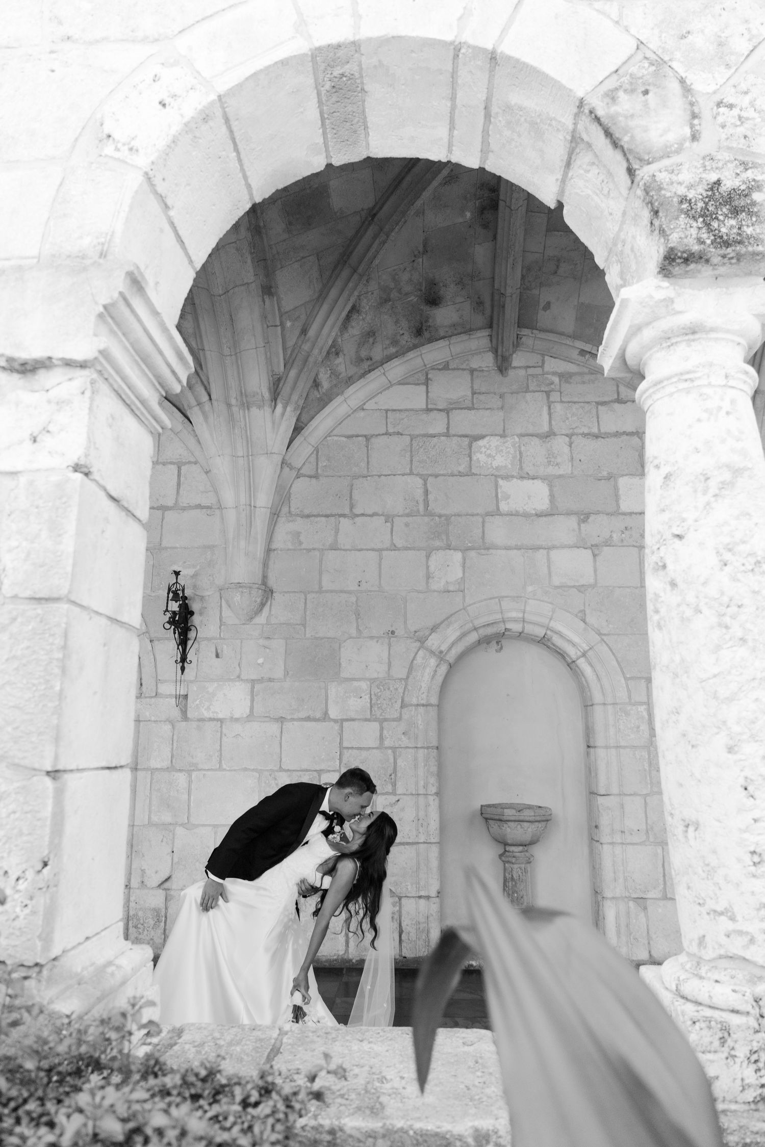 Black and white photo of bride and groom kissing under a stone arch at historic wedding venue.