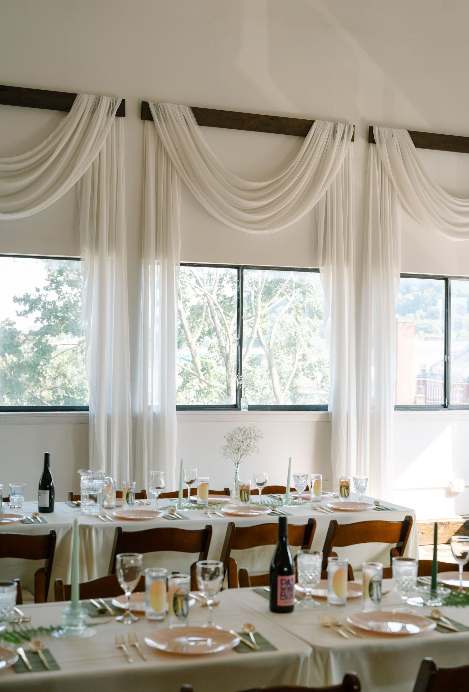 Wedding reception tables decorated with white linens and centerpieces near large windows.