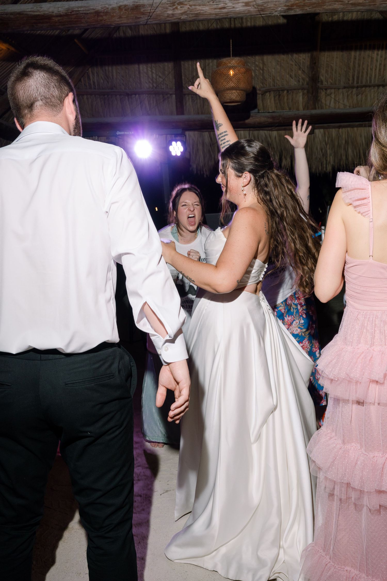 A group of people dancing and celebrating at a formal event, with a woman in a long white dress standing in the center of the frame.