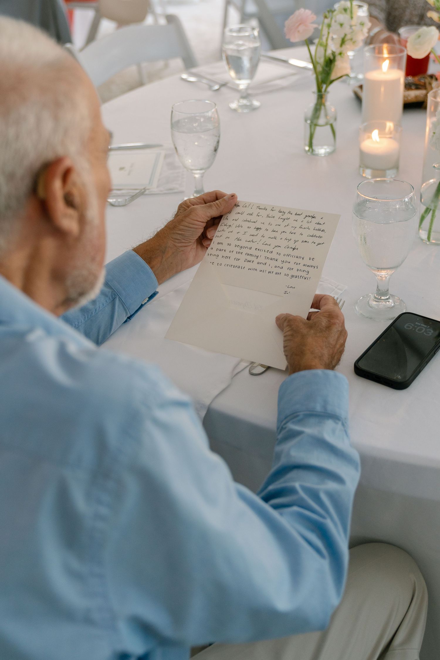 An elderly man is writing on a piece of paper on a table set for a formal dinner.