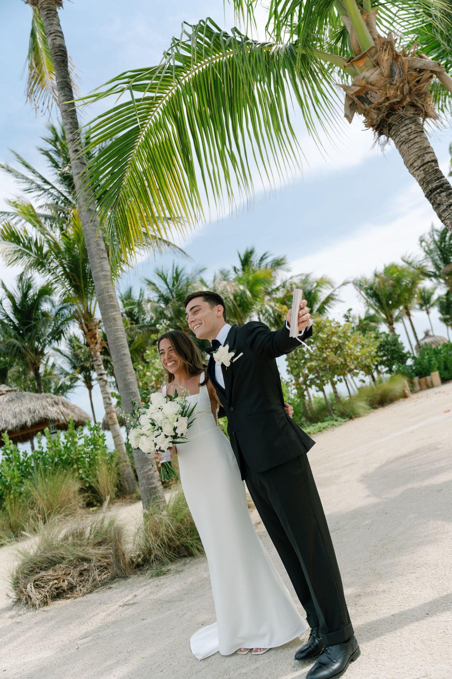 Bride and groom taking a selfie on a sandy beach surrounded by tall tropical palm trees.