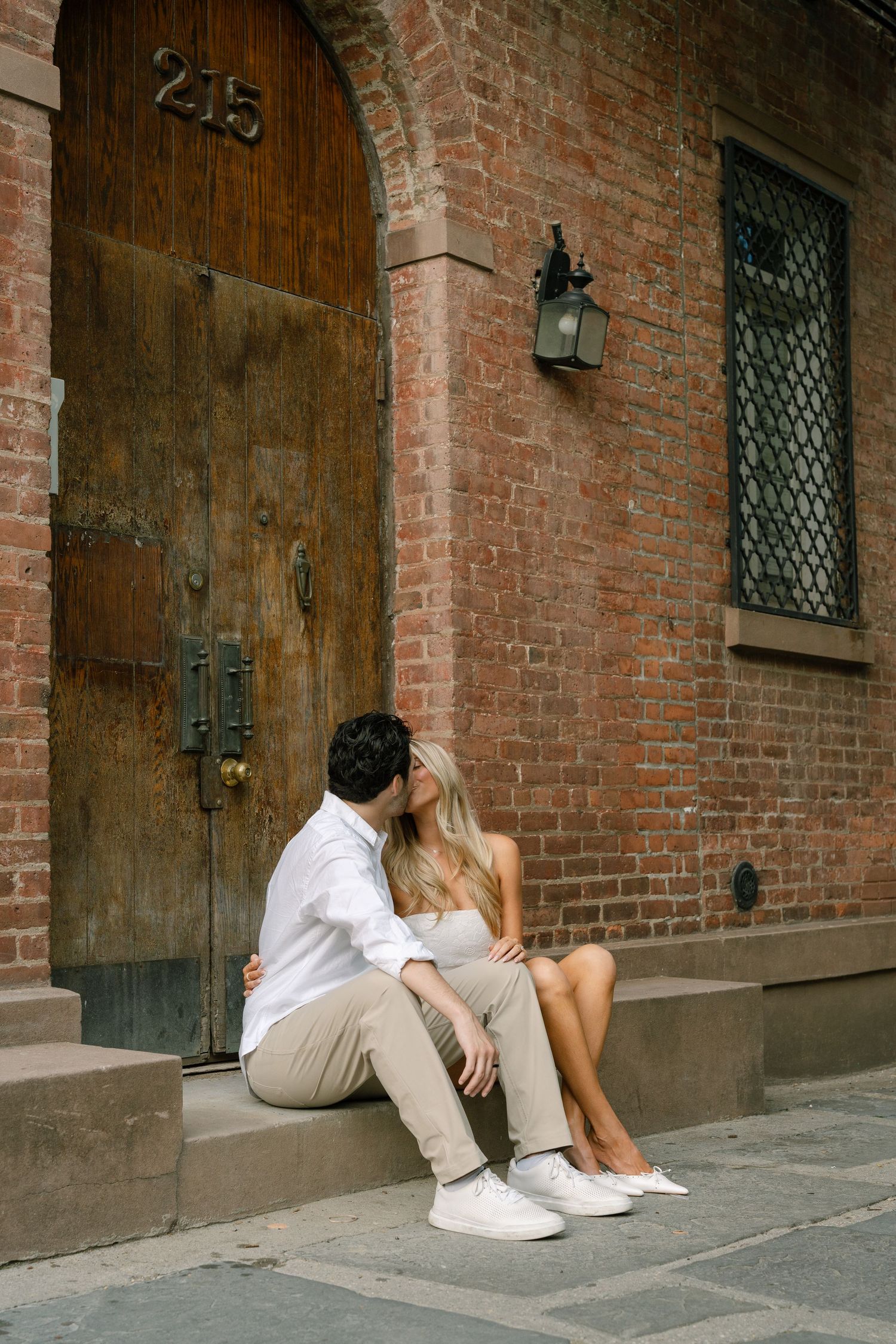 Couple sharing a romantic kiss on steps outside a rustic brick building with wooden door in NYC.