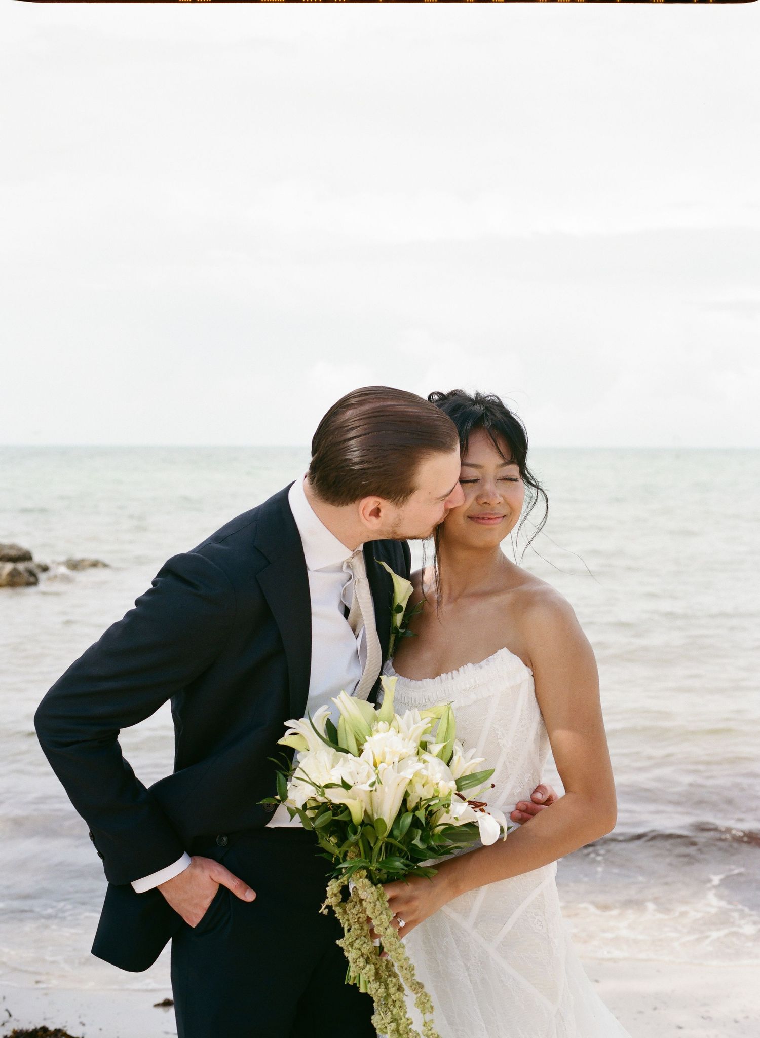 Groom in navy suit kisses bride in white strapless gown holding lily bouquet by the ocean shore.