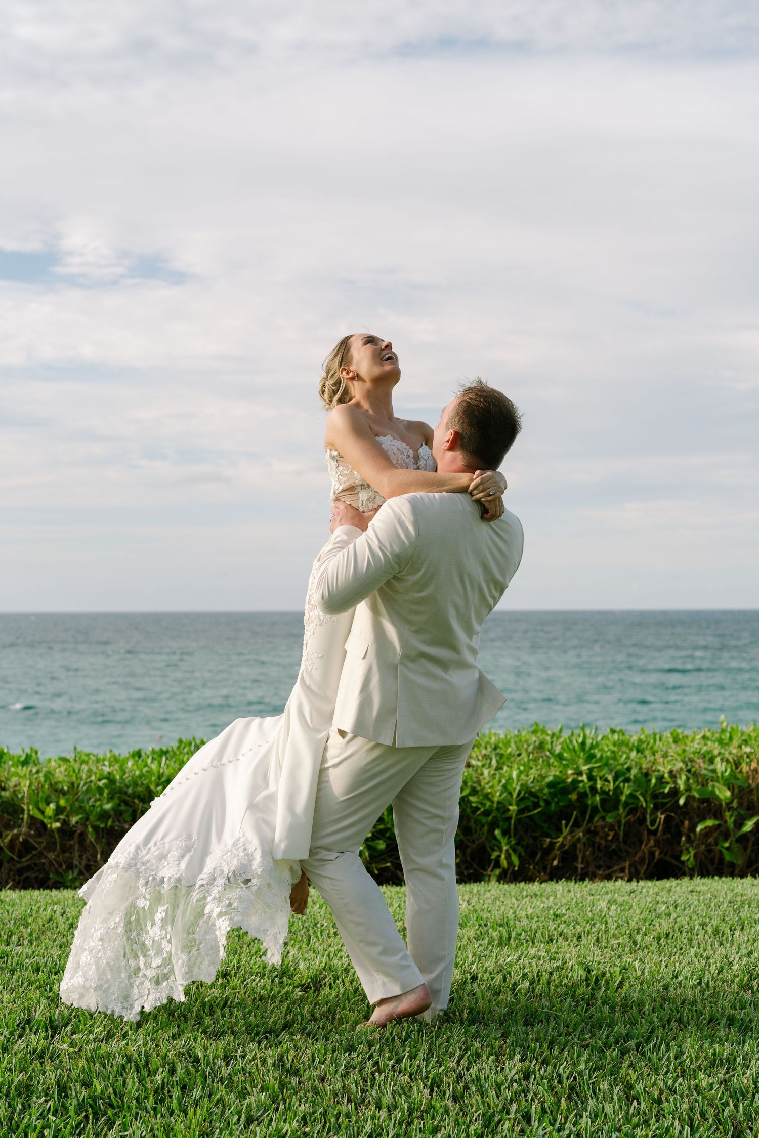 Groom lifting bride in white dress on green lawn with ocean backdrop at tropical wedding celebration.