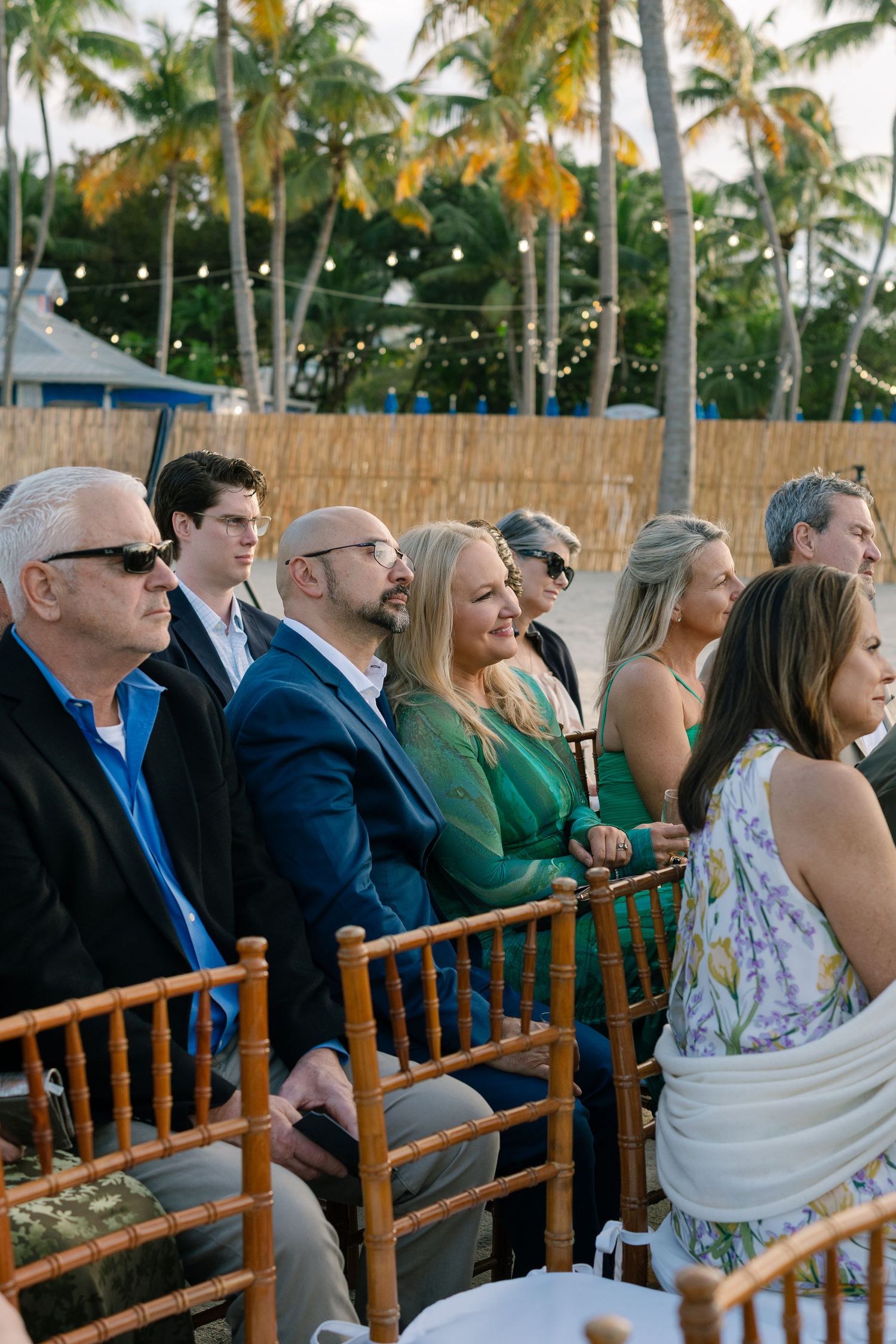 Wedding guests seated in wooden chairs under palm trees at an outdoor tropical ceremony.