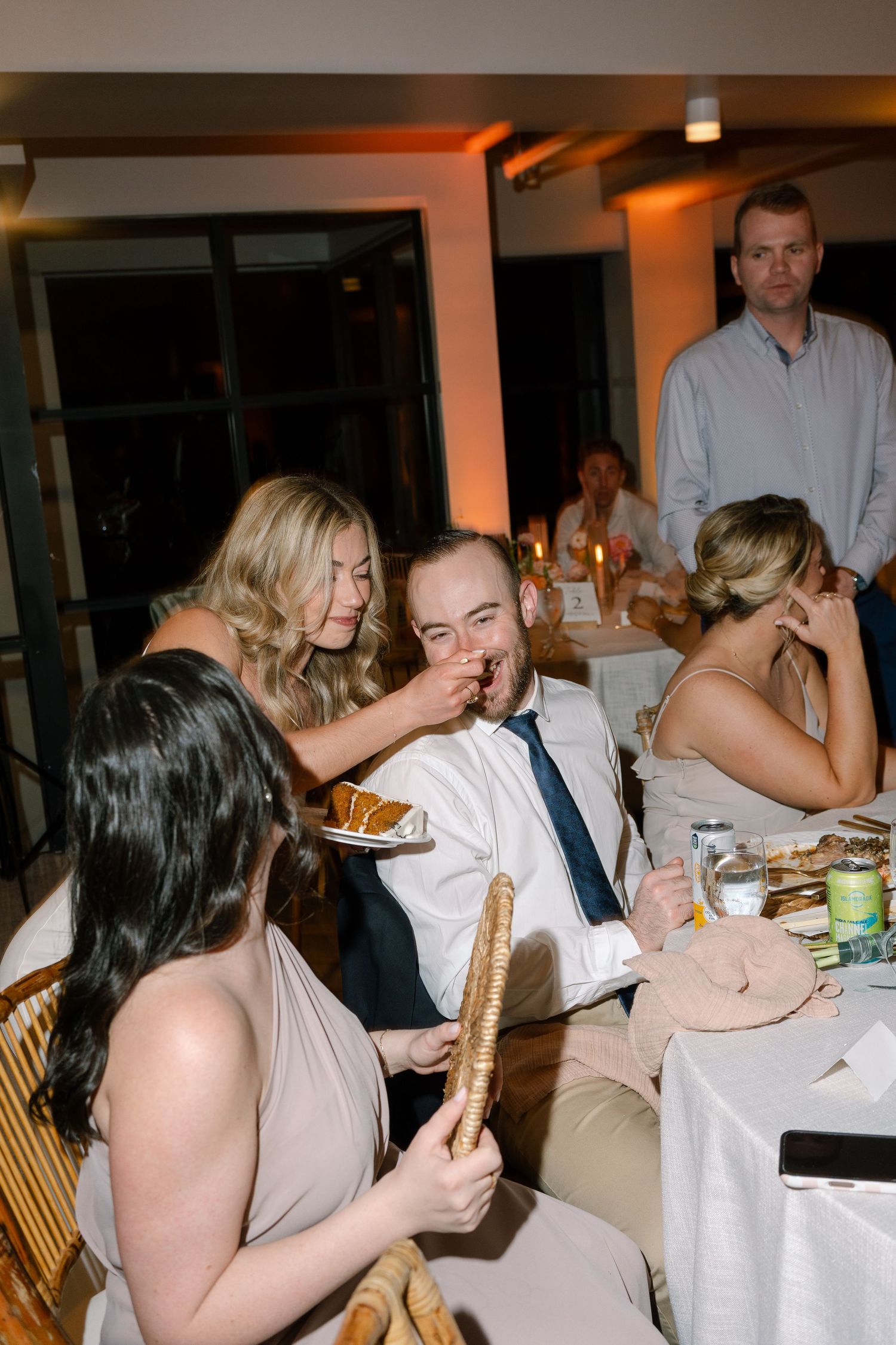 Lively wedding reception scene with guests sharing food and laughing around a decorated dining table.