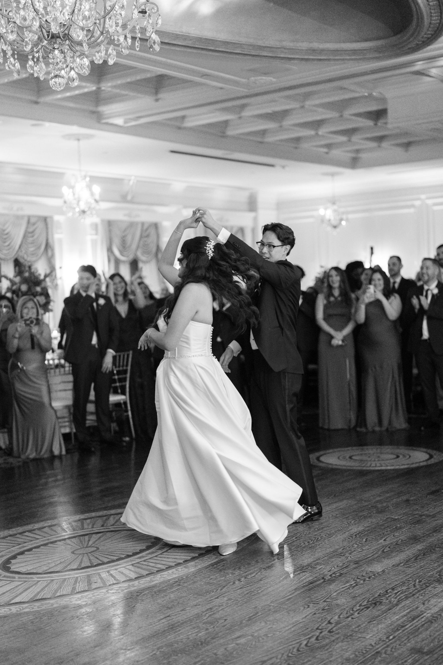 Wedding reception dance in a grand ballroom with couples twirling on a wooden dance floor.