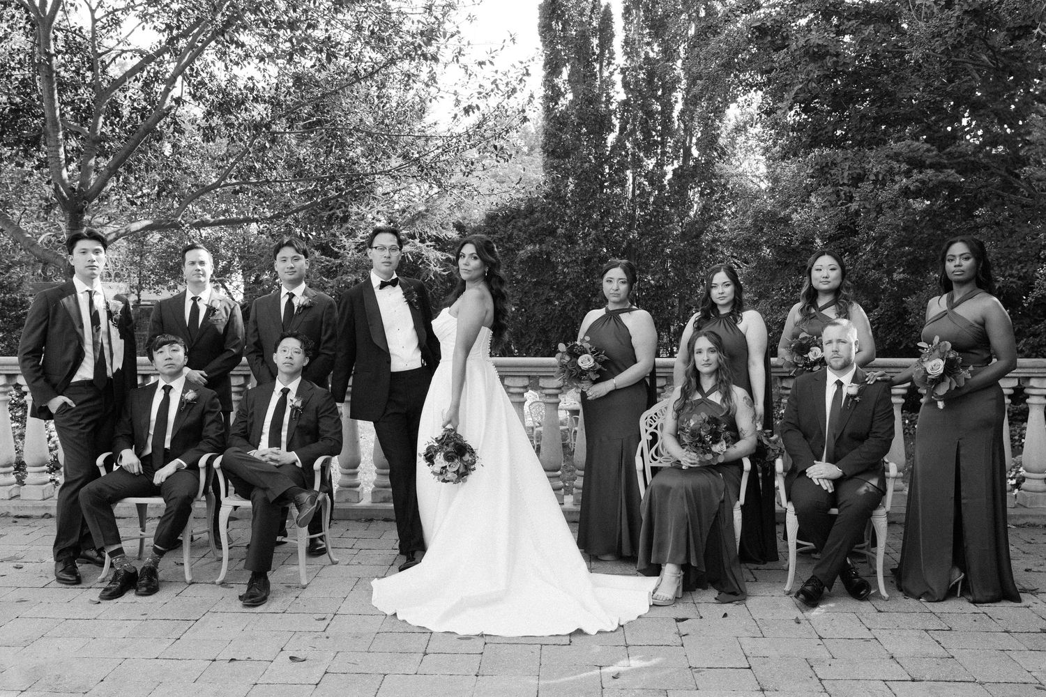 Large wedding party in formal attire arranged for group photos on outdoor stone steps.