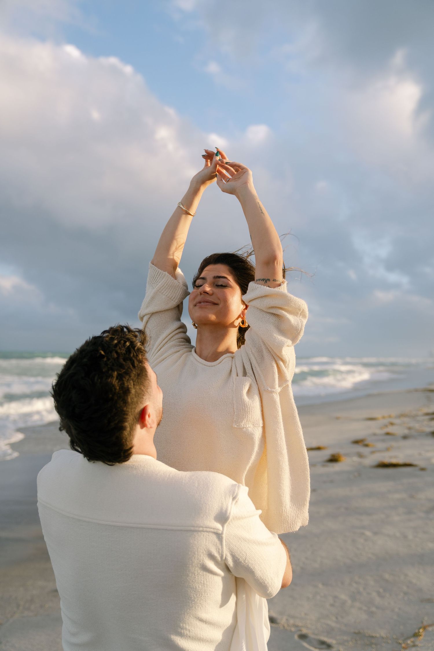 A couple in white outfits dance together on a beach at sunset with waves and cloudy skies in the background.