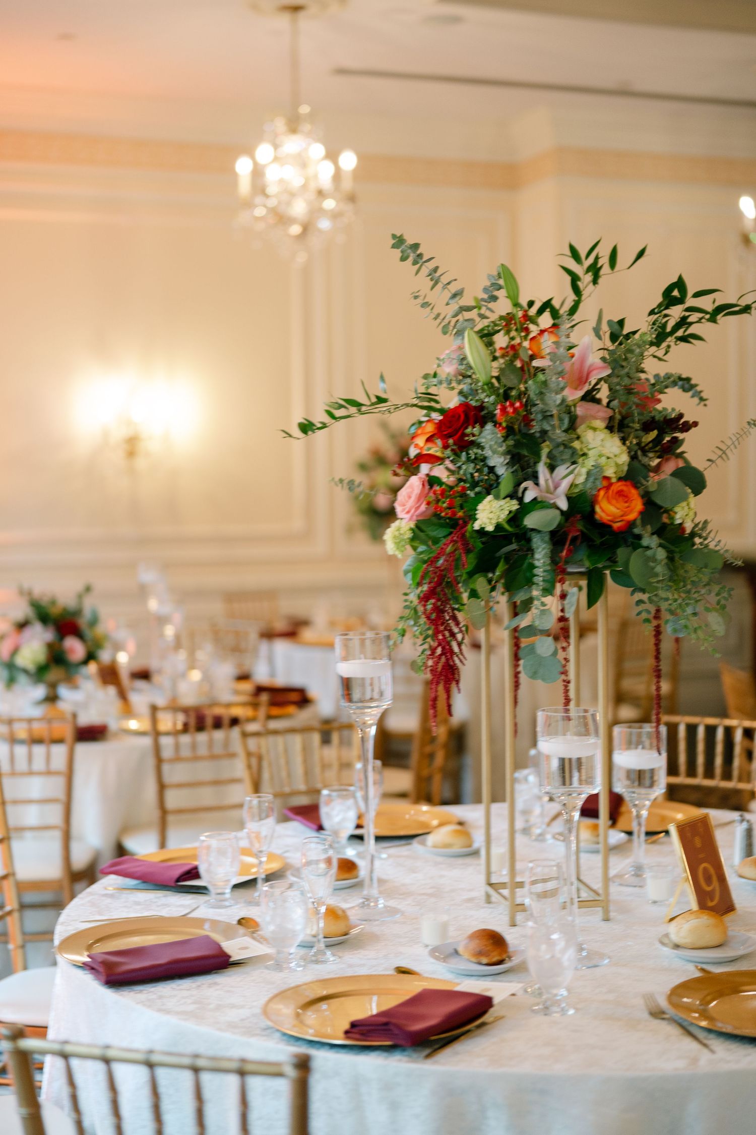 Elegant wedding reception tables decorated with tall floral centerpieces, burgundy napkins, and gold charger plates in a bright ballroom.