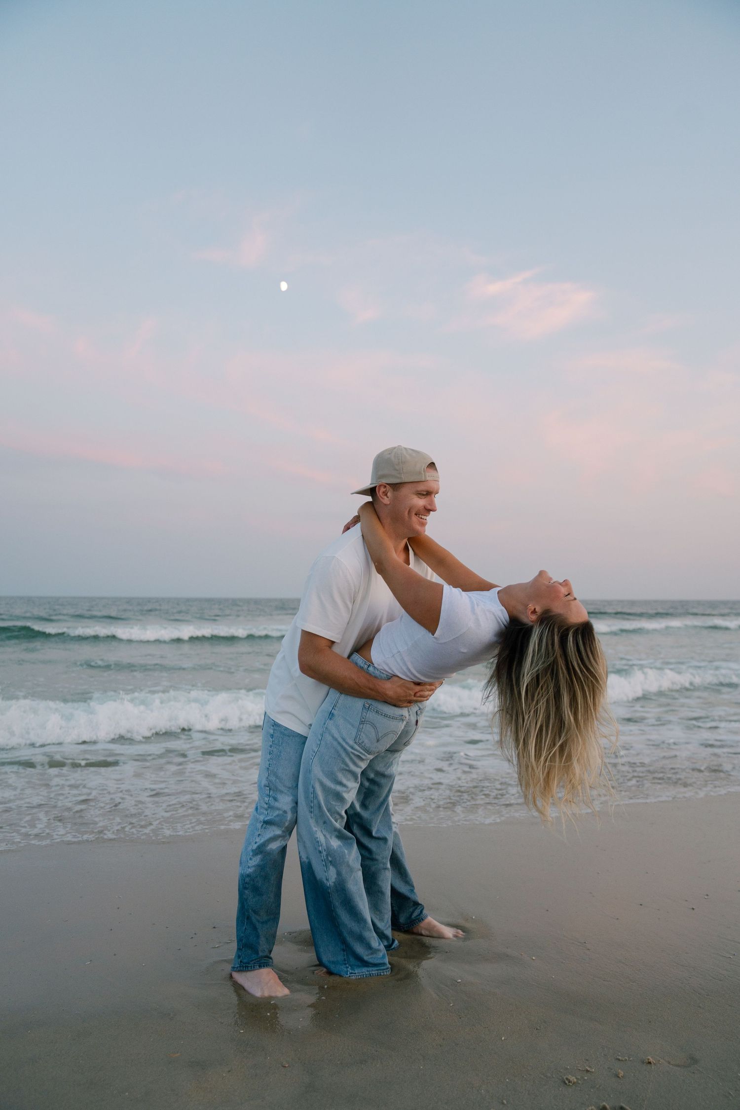 Romantic couple embraces on a beach at sunset with rolling waves and pastel pink sky in the background.