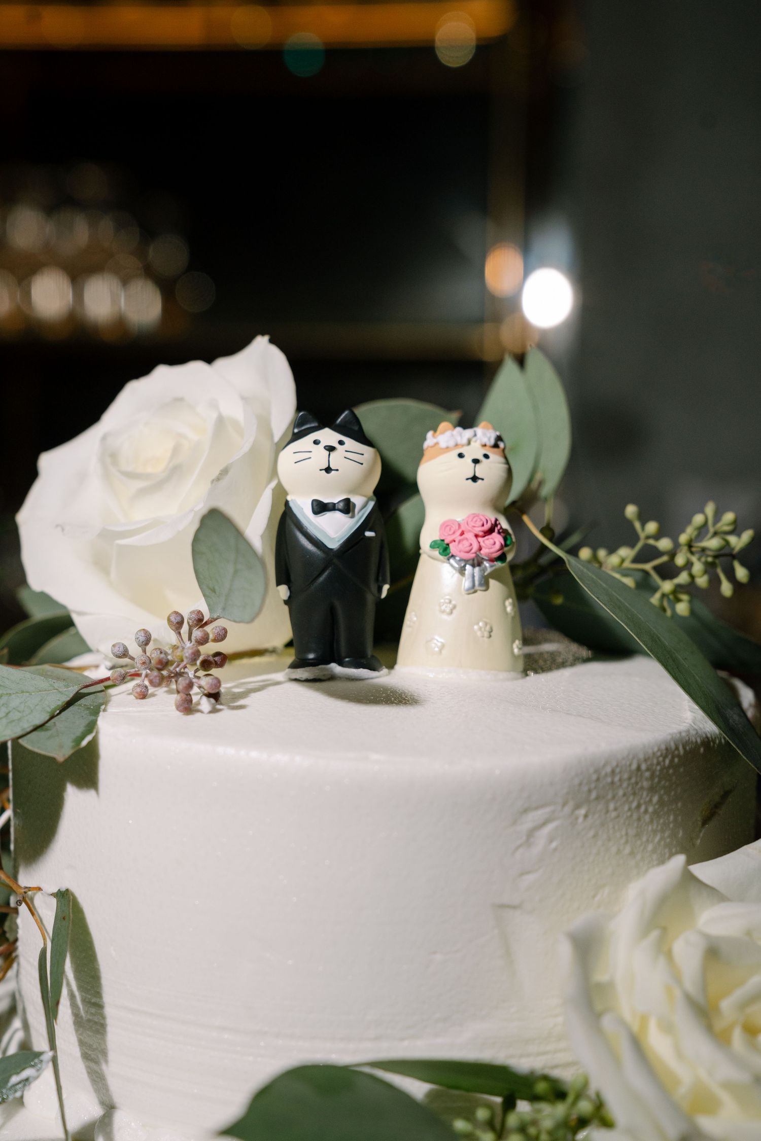 Adorable cat-shaped wedding cake toppers sit atop a white frosted cake with eucalyptus leaves and roses.