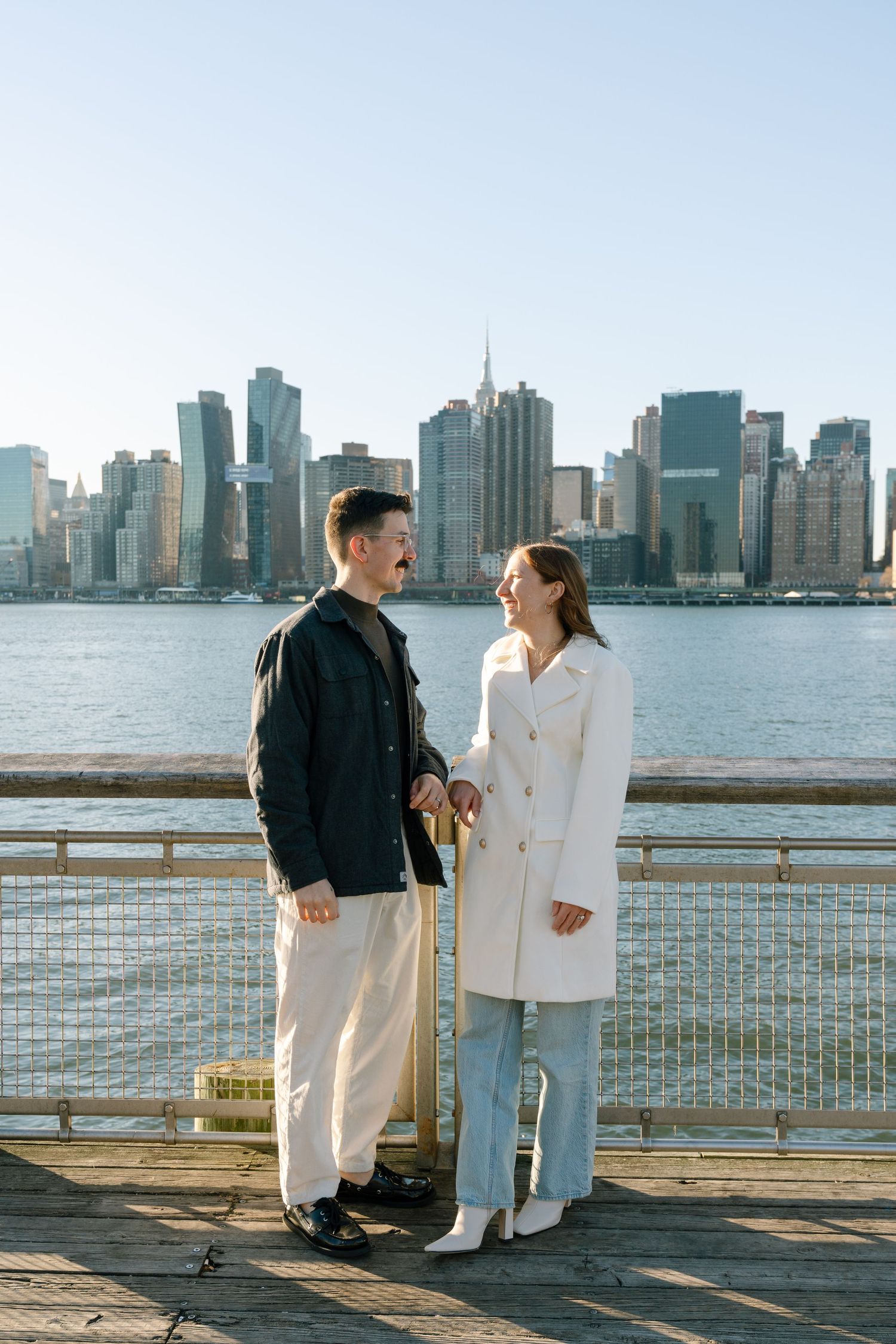 A couple stands together on a waterfront pier with the Manhattan skyline visible across the water.