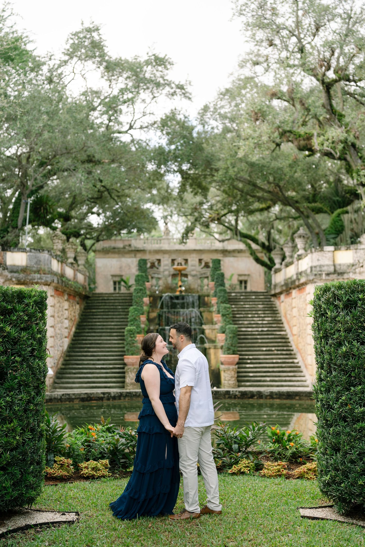 A couple stands together on a garden path surrounded by stone steps and lush greenery.