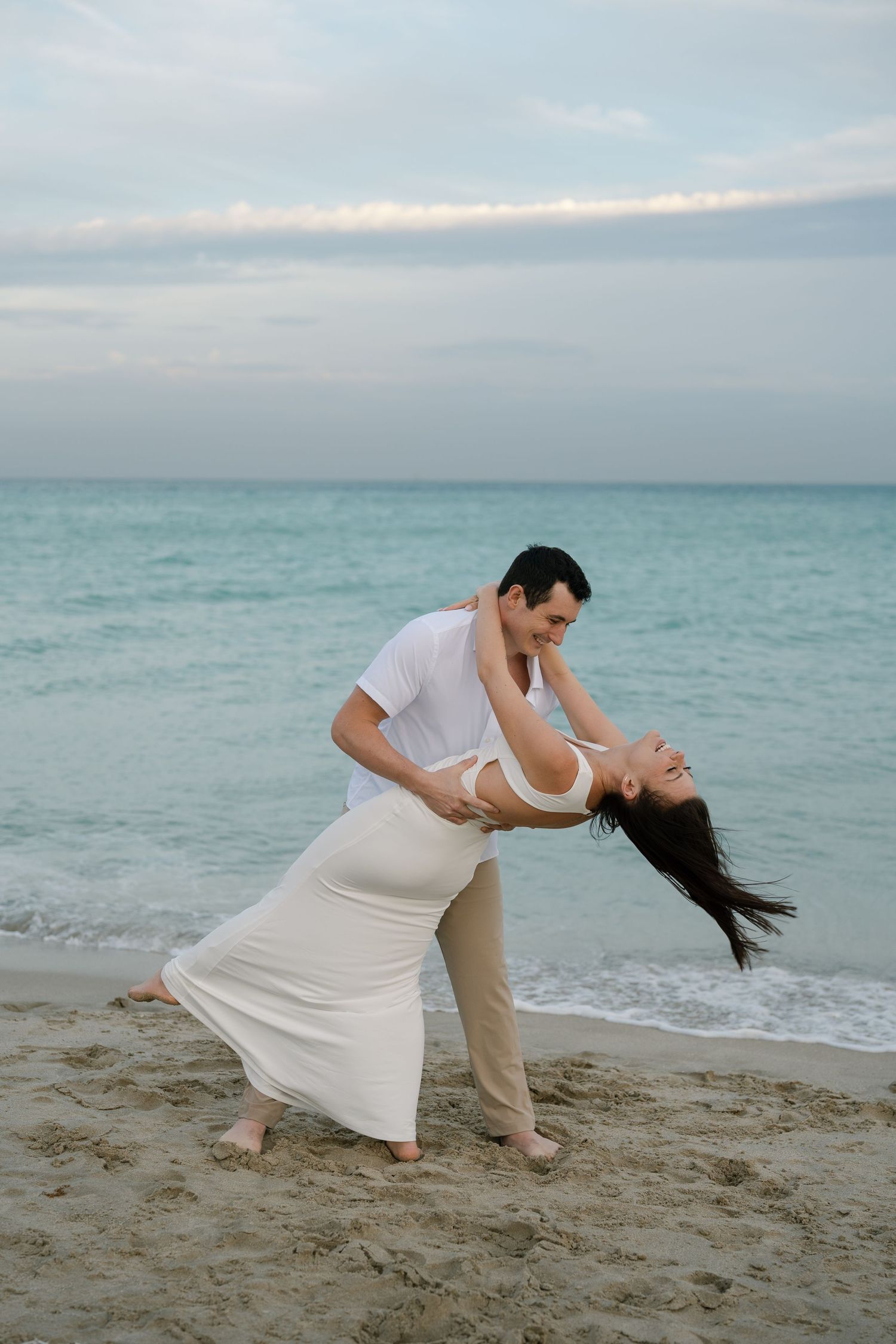 A couple performs a romantic dance dip pose on a sandy beach with gentle waves in the background.